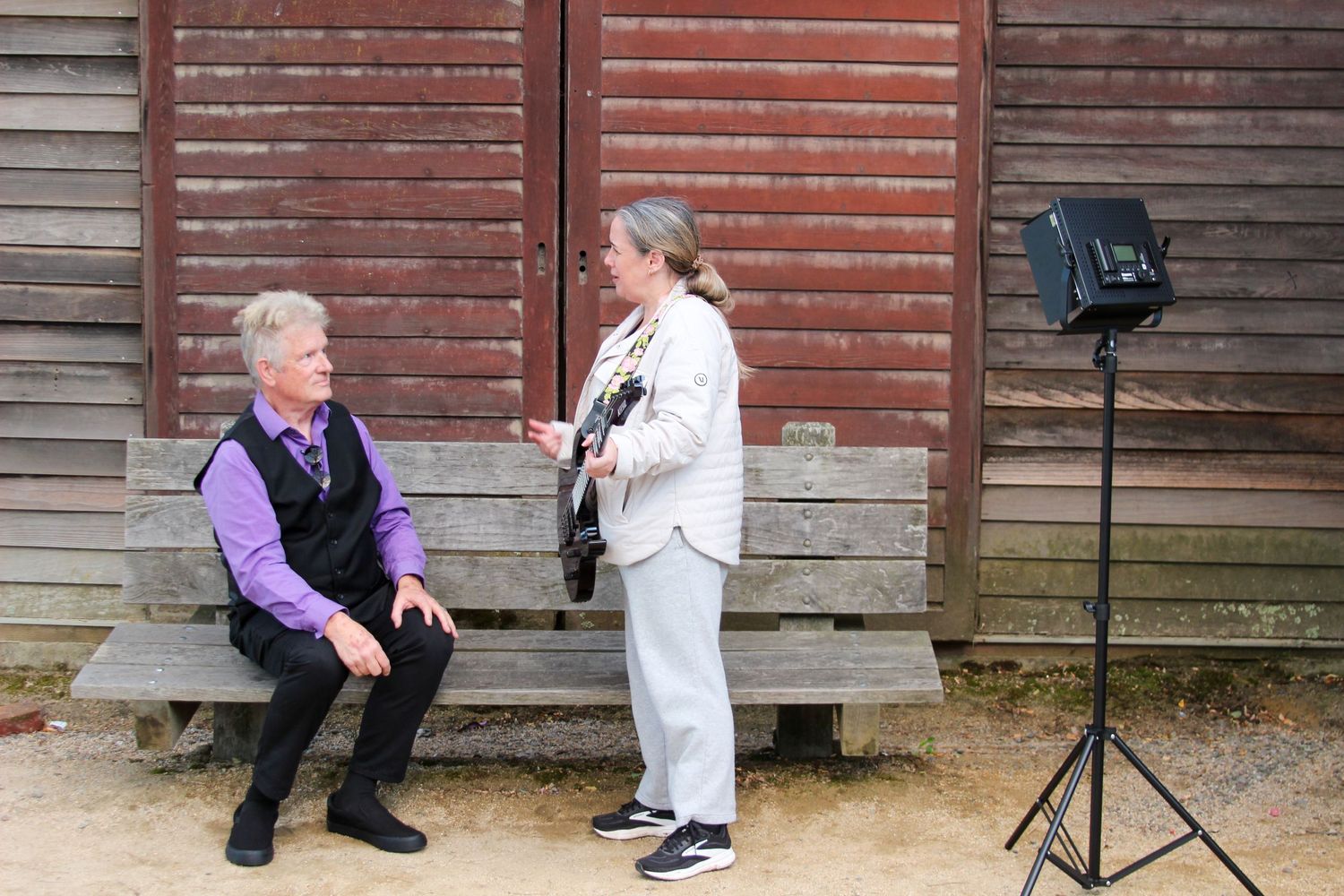 Two musicians outdoors near rustic barn, one seated on bench, other standing with guitar, professional lighting setup nearby.