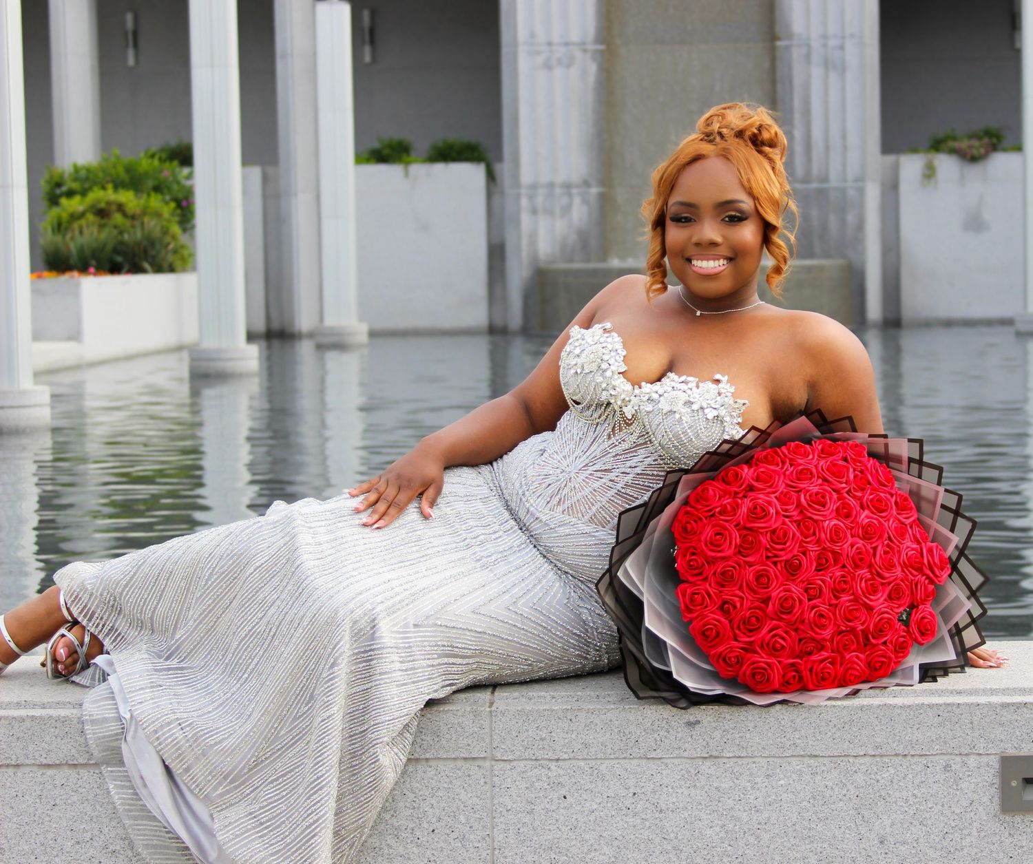 A woman in a white strapless gown sits elegantly holding a large bouquet of red roses near a reflective pool.
