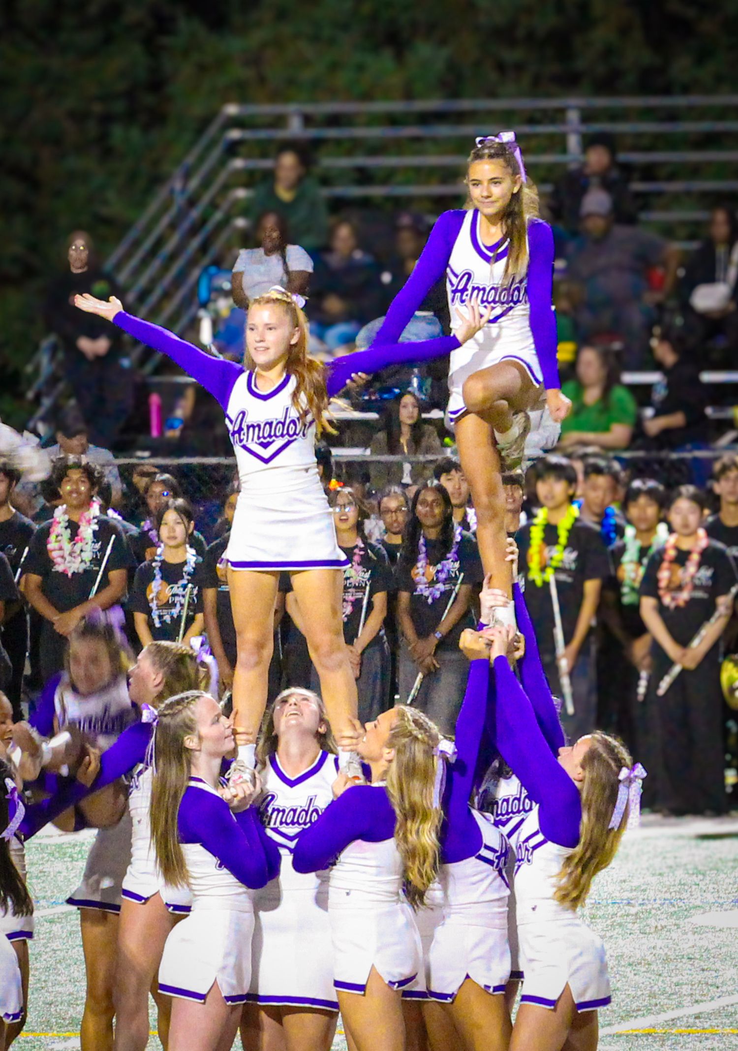 Amador cheerleaders perform a stunt pyramid in purple and white uniforms at a night football game with crowd in background.