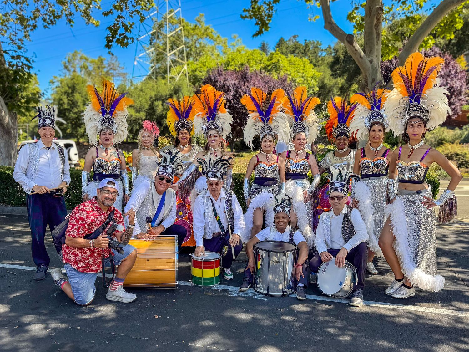 Colorful samba dancers in feathered headdresses pose with drummers outdoors at a festive carnival performance.