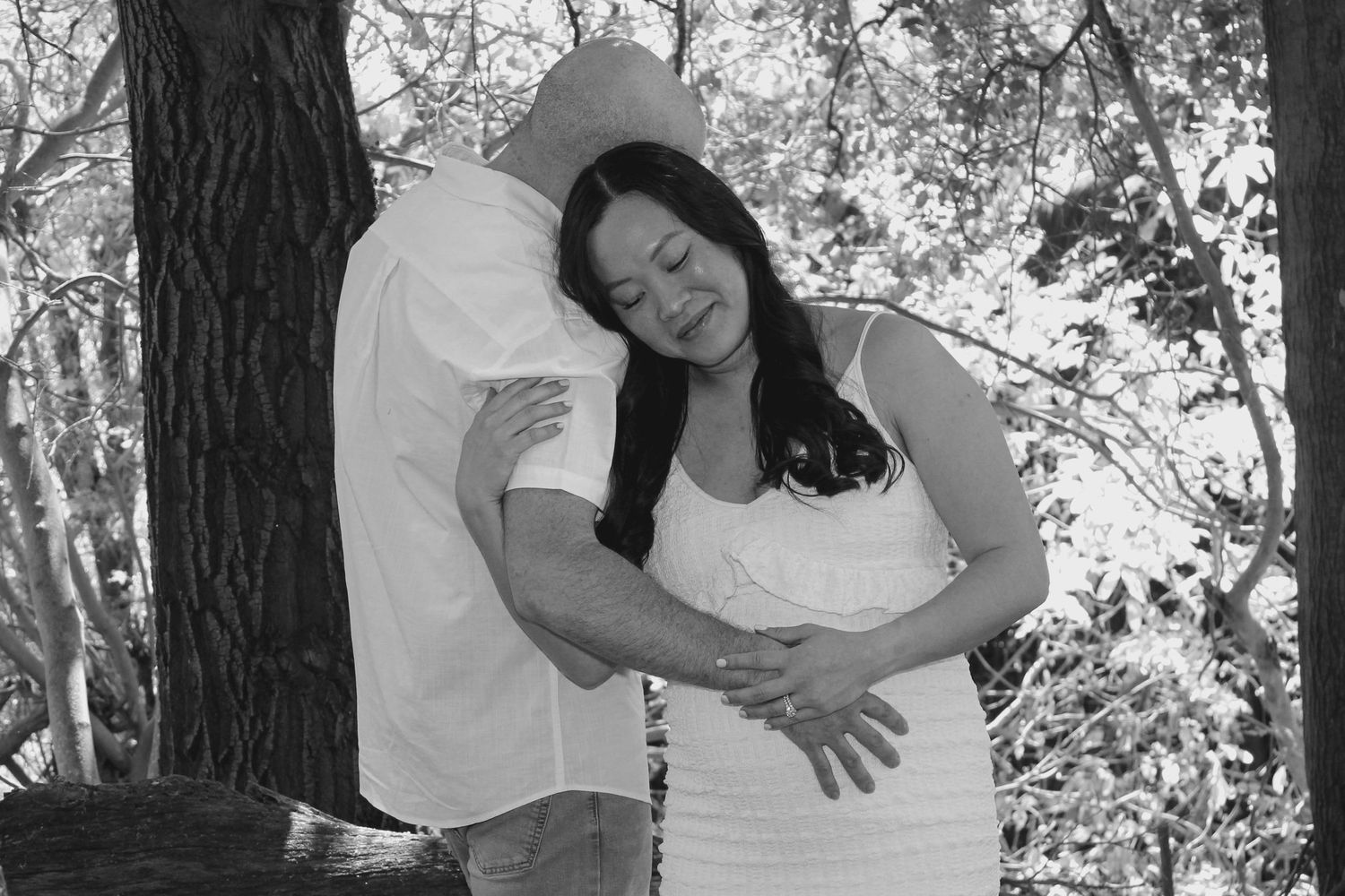 Pregnant woman in white dress embraced by partner among trees in a black and white maternity photo shoot.