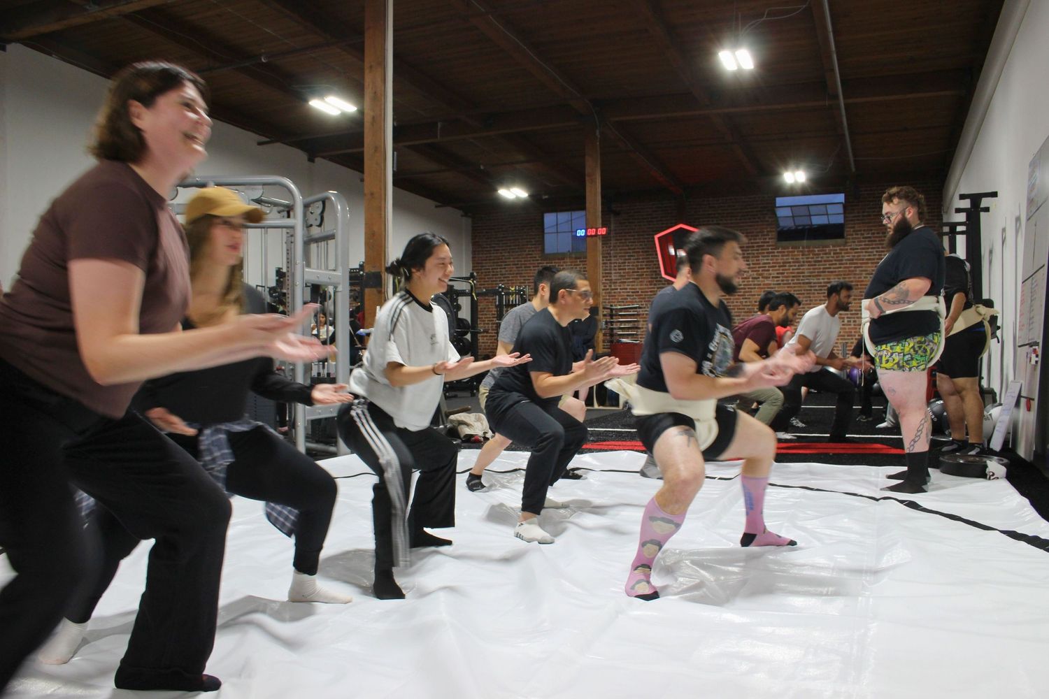 Group fitness class performing squats together in a modern gym with equipment and red accents in background.