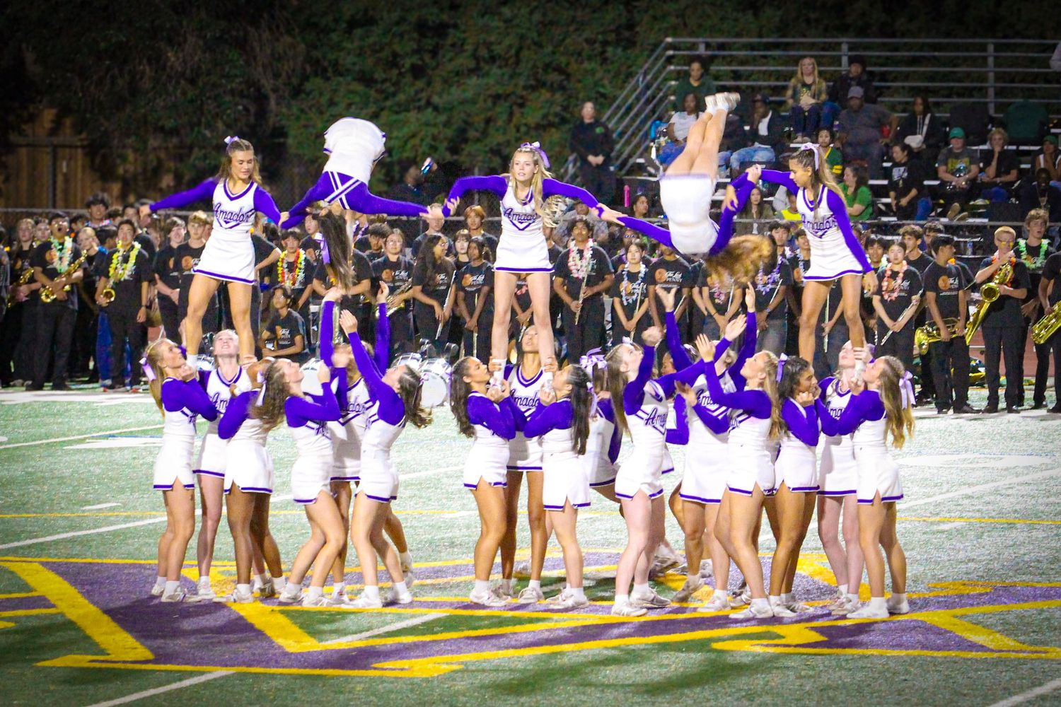 Cheerleaders in purple and white uniforms perform a pyramid stunt on a football field at a night game.
