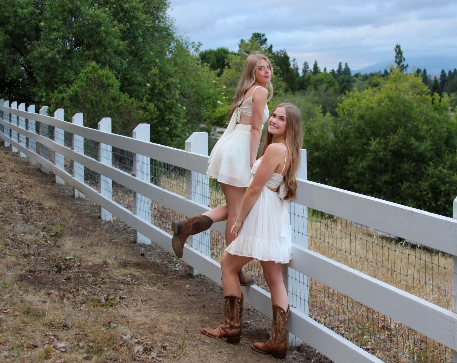 Two young women in white dresses and cowboy boots pose by a white wooden fence in a rural countryside setting.