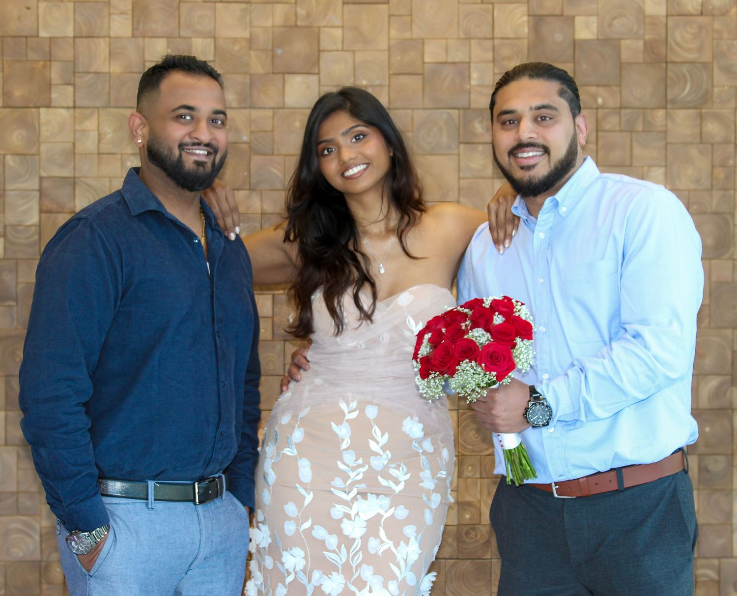 Bride in lace wedding dress holding red bouquet poses with two men in casual attire against wood wall.