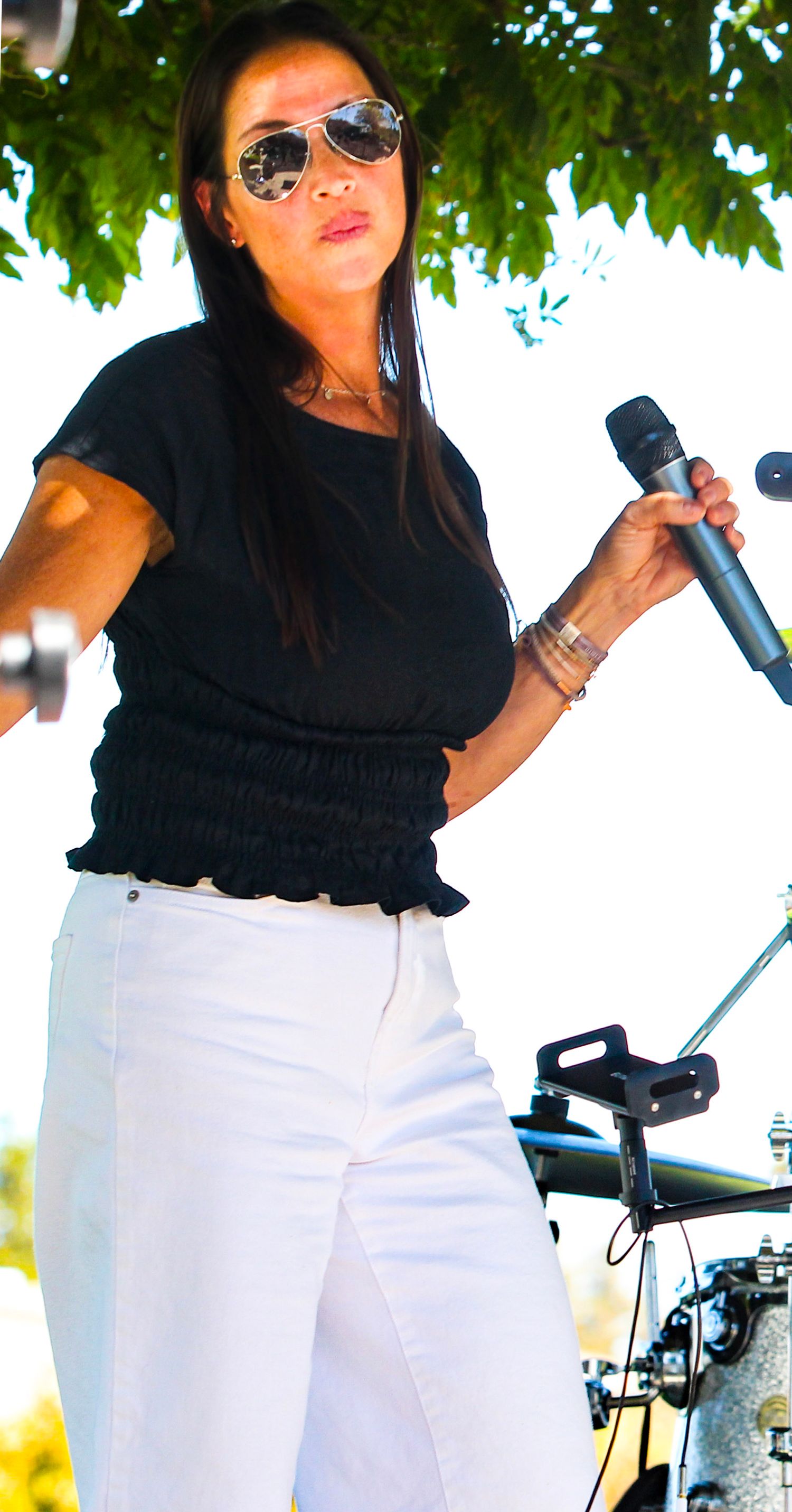 Female singer performing outdoors in white pants and black top, holding microphone while wearing aviator sunglasses.