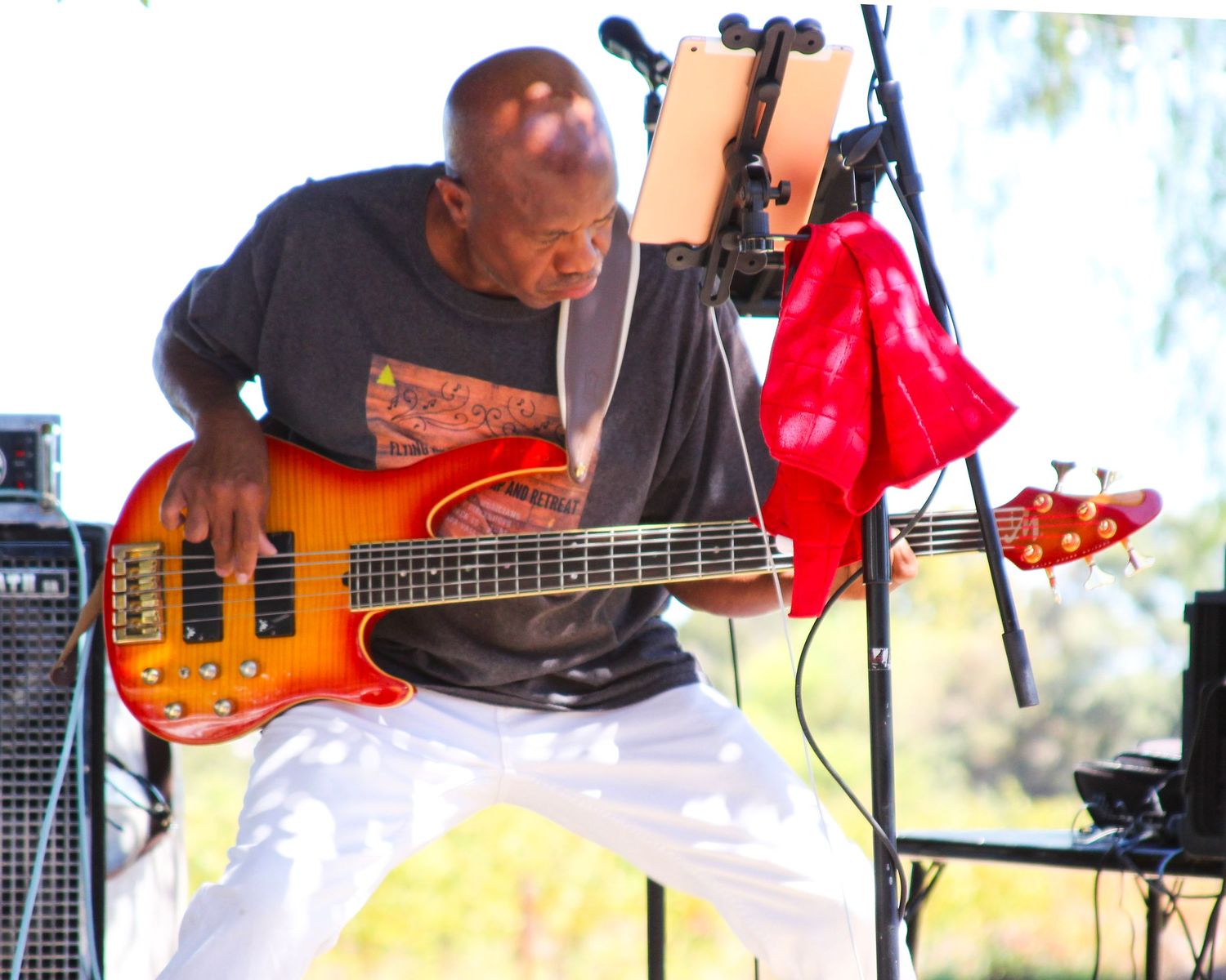 Bald musician passionately playing a sunburst bass guitar on stage, wearing white pants and gray t-shirt.