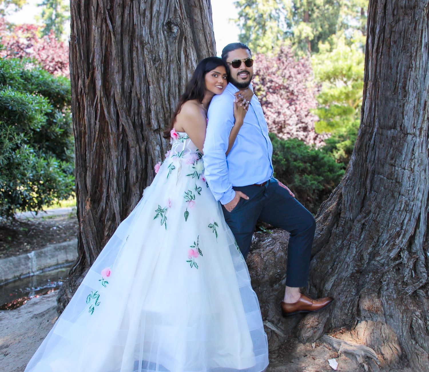 Couple posing by large tree trunks, woman in white floral ball gown, man in blue shirt and navy pants, outdoor garden setting.