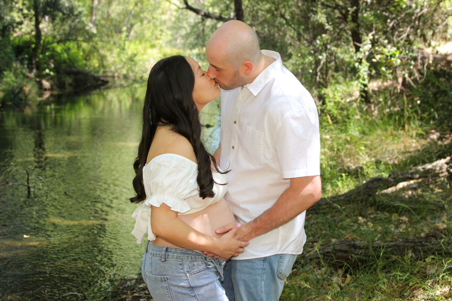 A couple shares a romantic kiss near a serene river surrounded by lush green trees outdoors.