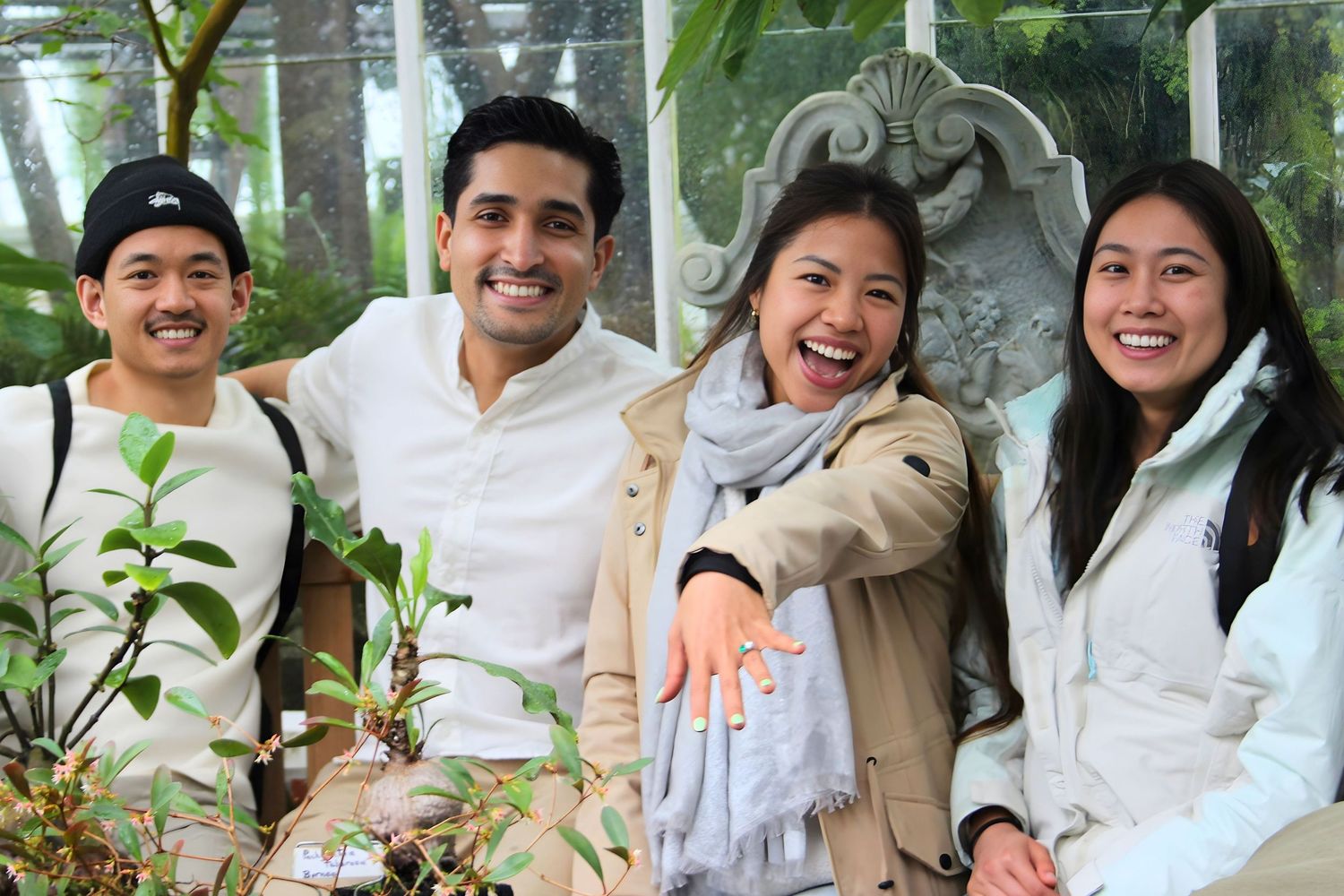 Four smiling friends posing together among green plants in a greenhouse setting.