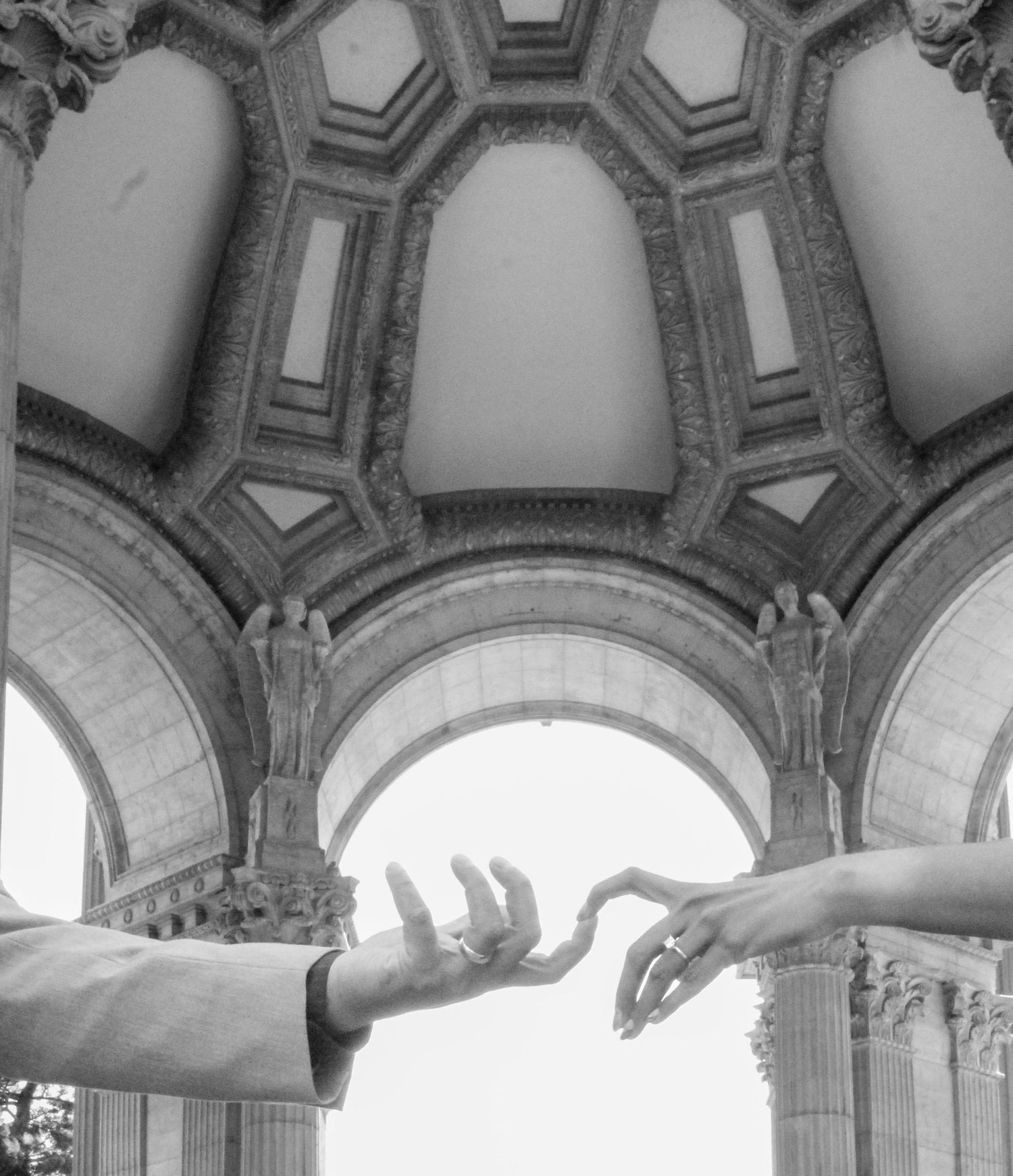 Black and white photo of two hands almost touching beneath ornate arched architecture, reminiscent of Michelangelo's Creation of Adam.