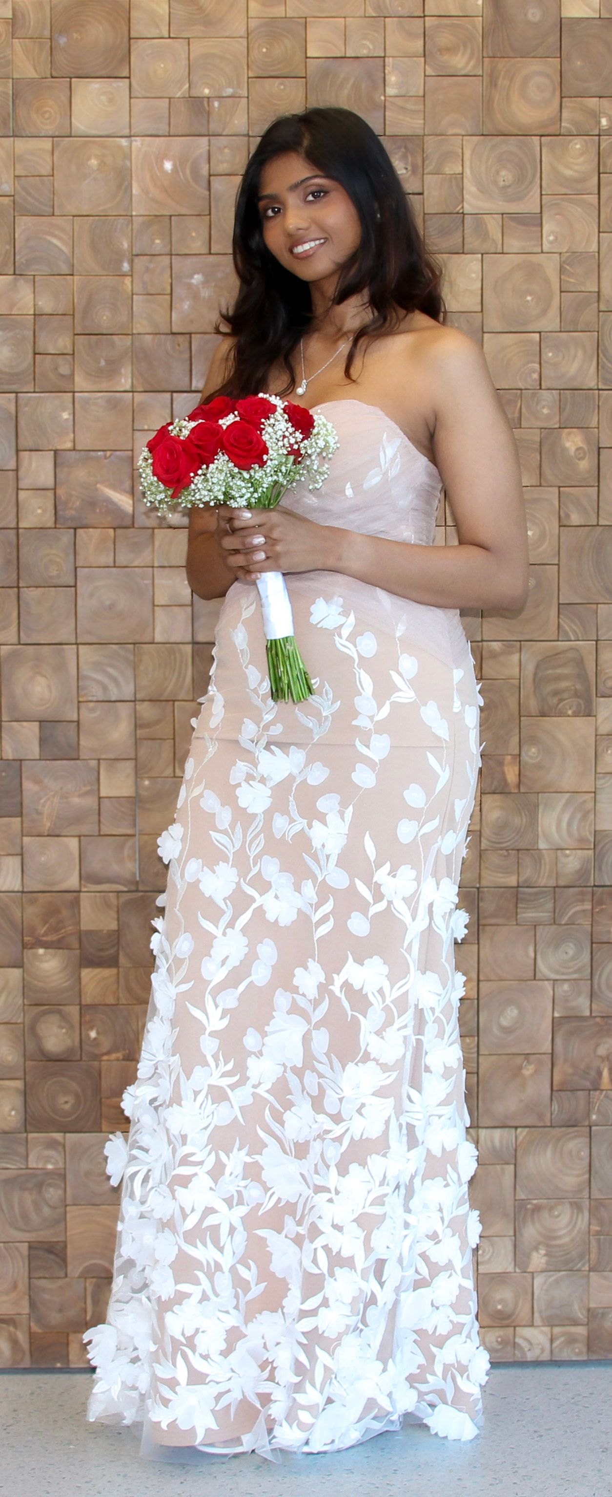 Woman in white floral strapless gown holding red rose bouquet with baby's breath, smiling at wedding ceremony.
