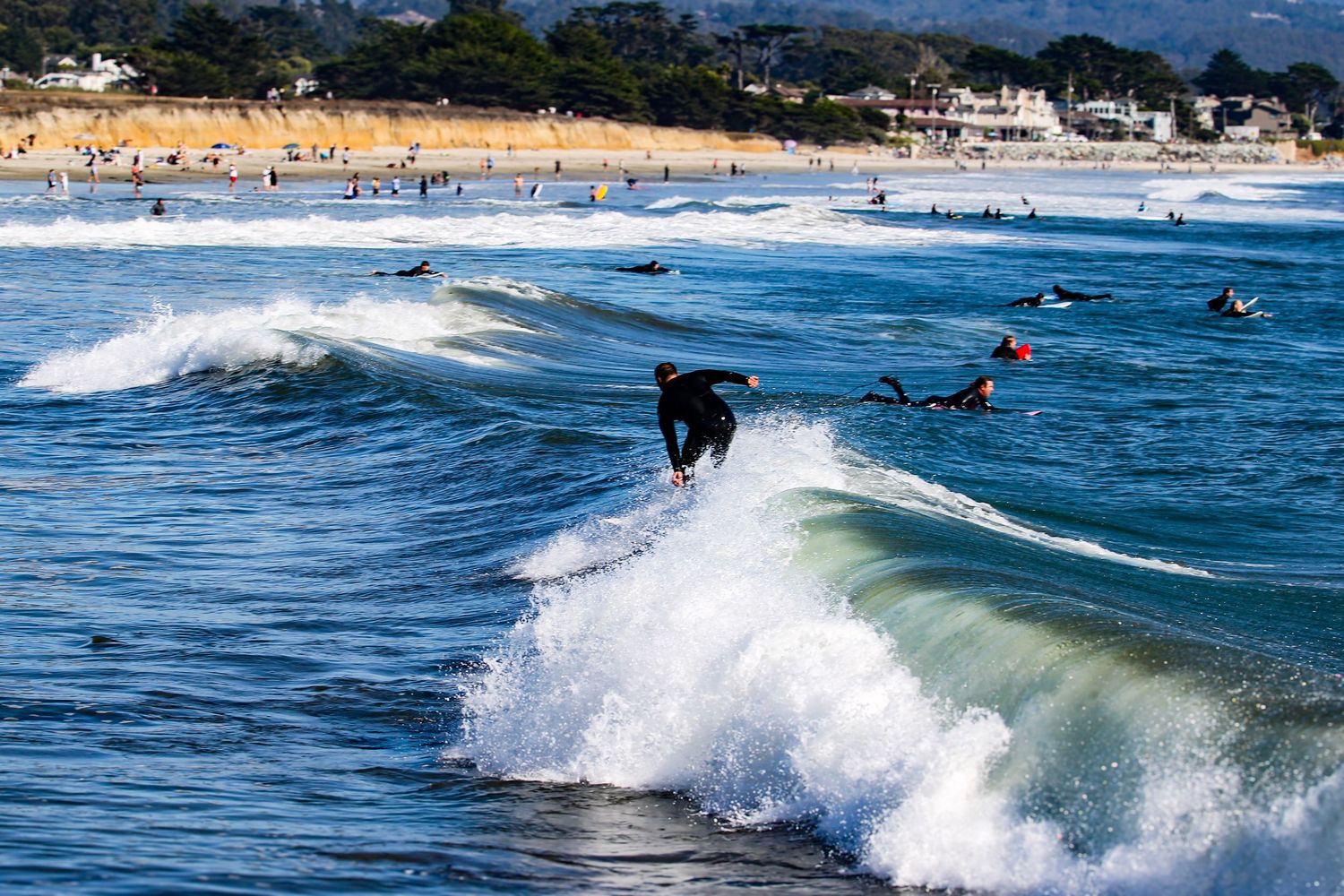 Surfers riding ocean waves at a sunny beach with sandy shores and spectators visible in the background.