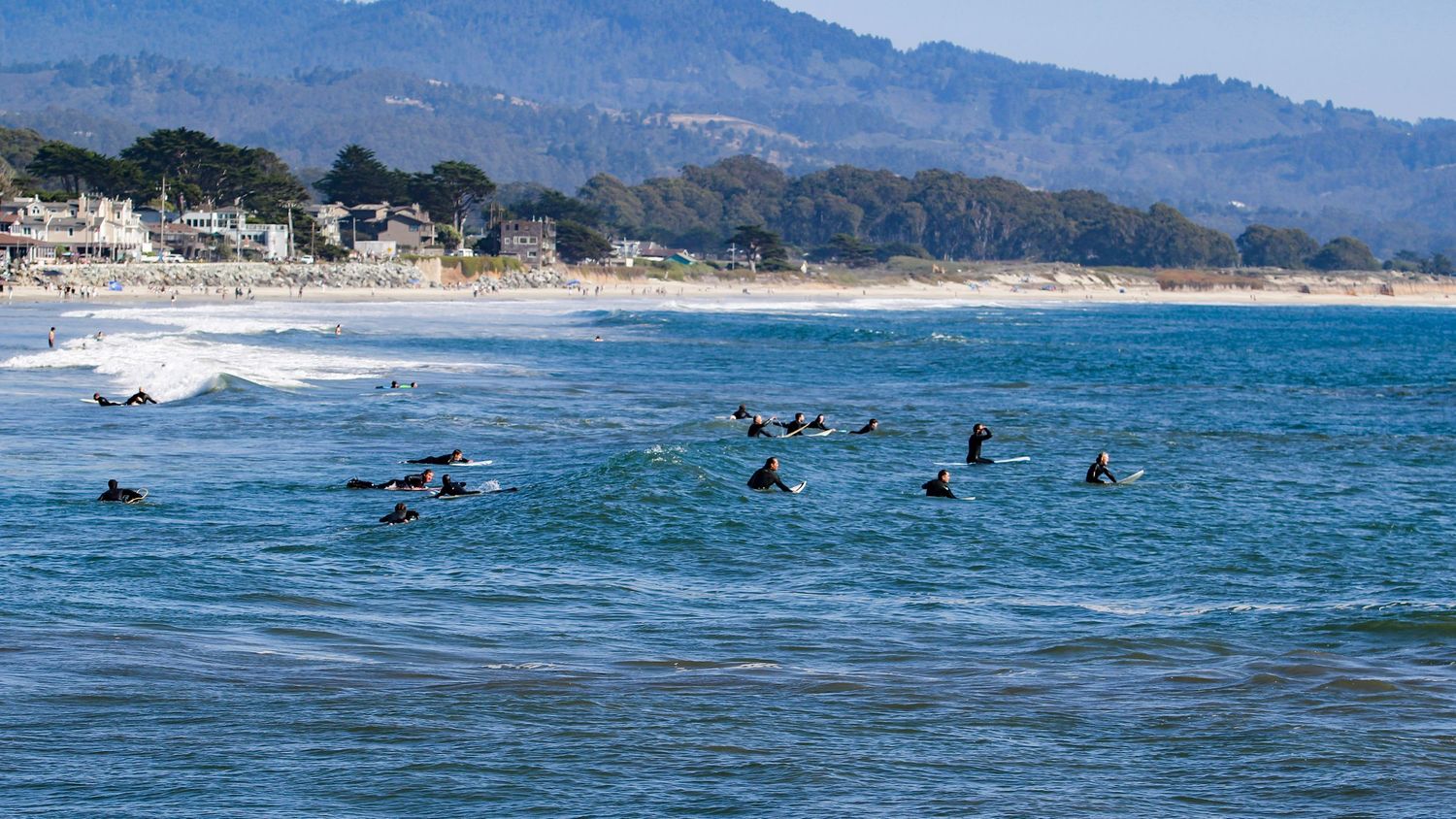 Surfers waiting for waves in the Pacific Ocean near a California beach town with mountains in the background.