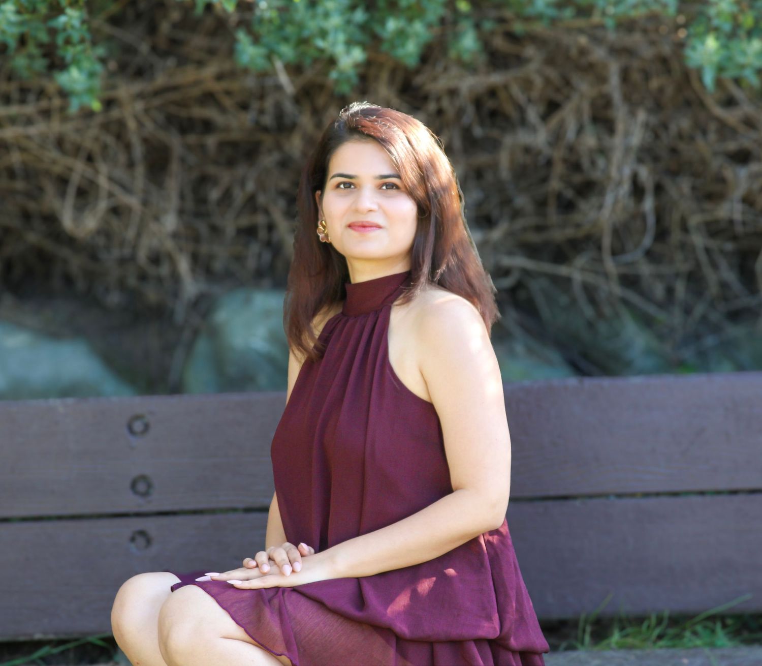 Young woman in burgundy halter dress sitting gracefully on a park bench with lush green foliage background.