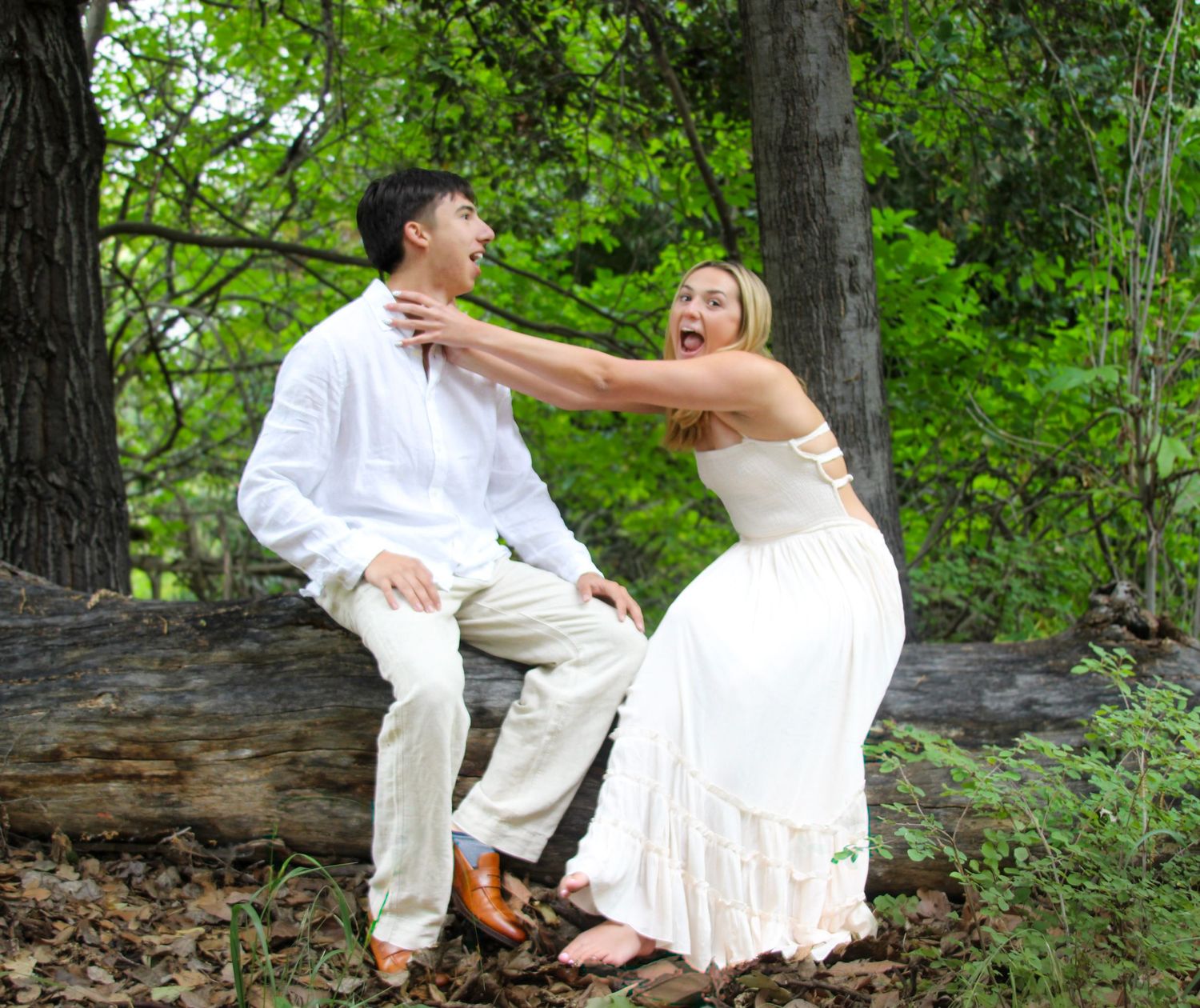 A joyful bride in a white dress playfully grabs her groom sitting on a log in a lush green forest setting.