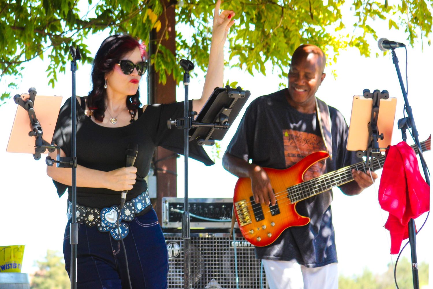 Female vocalist and bassist performing outdoors at live music event under green trees on sunny day.
