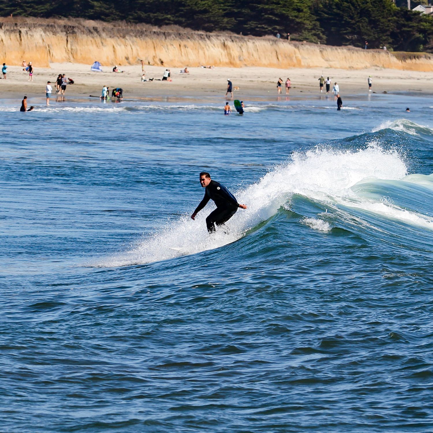 A surfer in a black wetsuit rides a wave at a sandy beach with people wading in the background.
