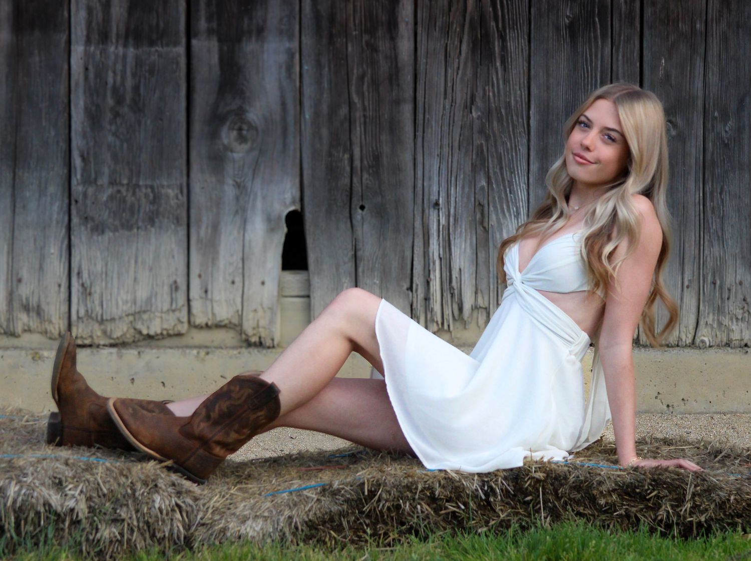 Young blonde woman in white dress and brown cowboy boots sitting against rustic wooden barn wall.