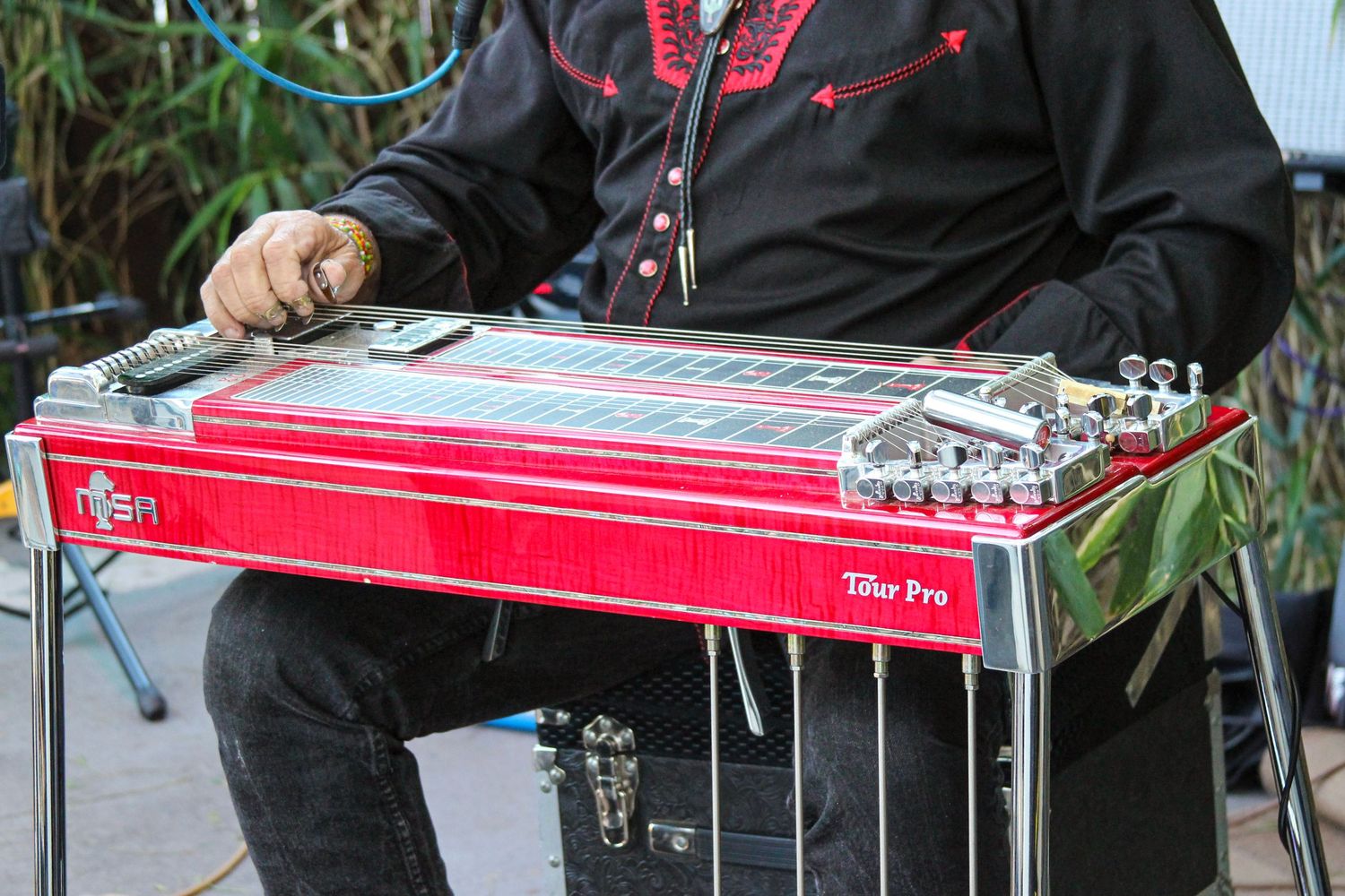Musician playing a red Tour Pro pedal steel guitar outdoors, wearing a black embroidered shirt.