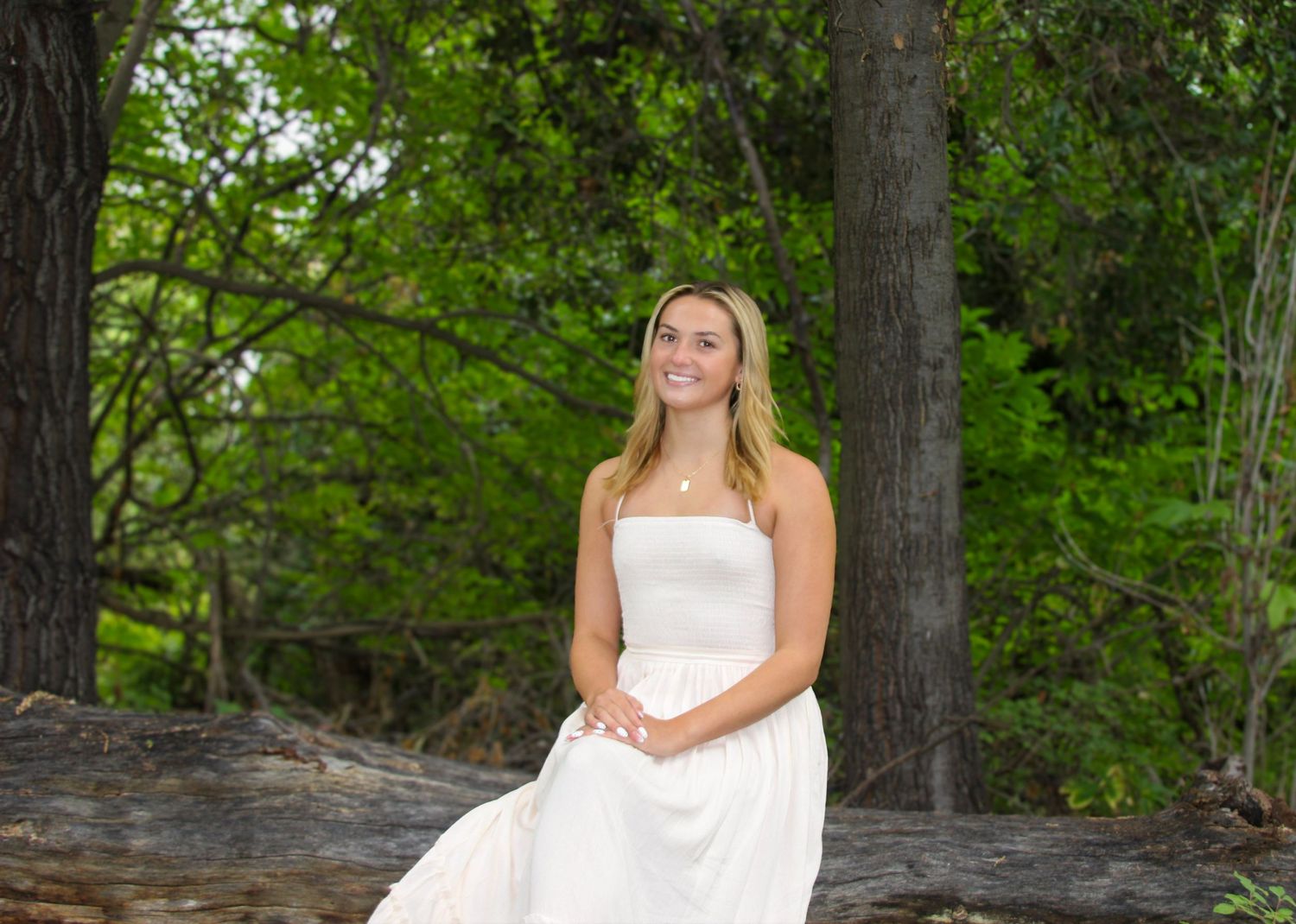 Young woman in white dress sitting on a log surrounded by lush green trees outdoors.