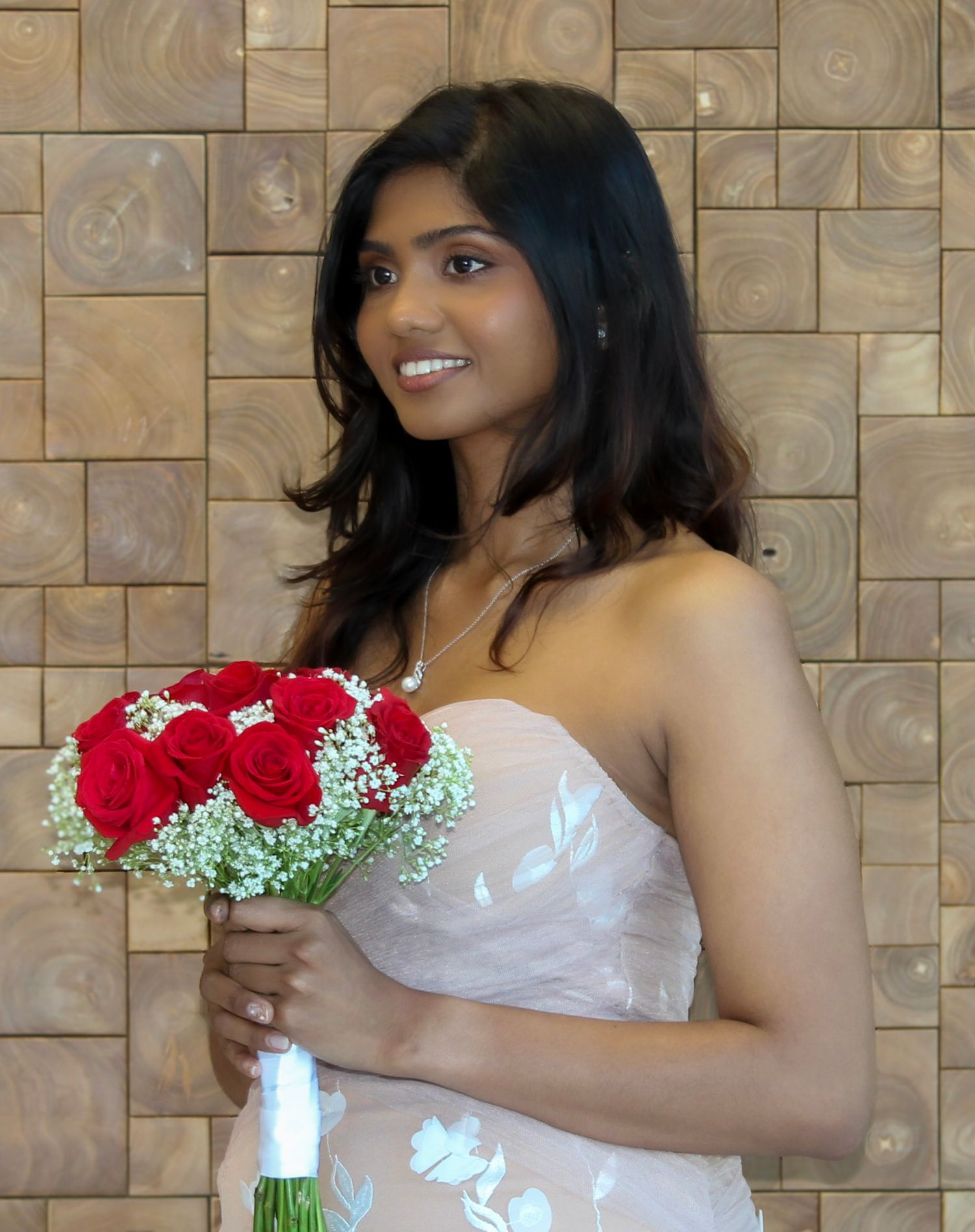 Young woman in strapless white gown holding red rose bouquet with baby's breath, smiling elegantly at a formal event.