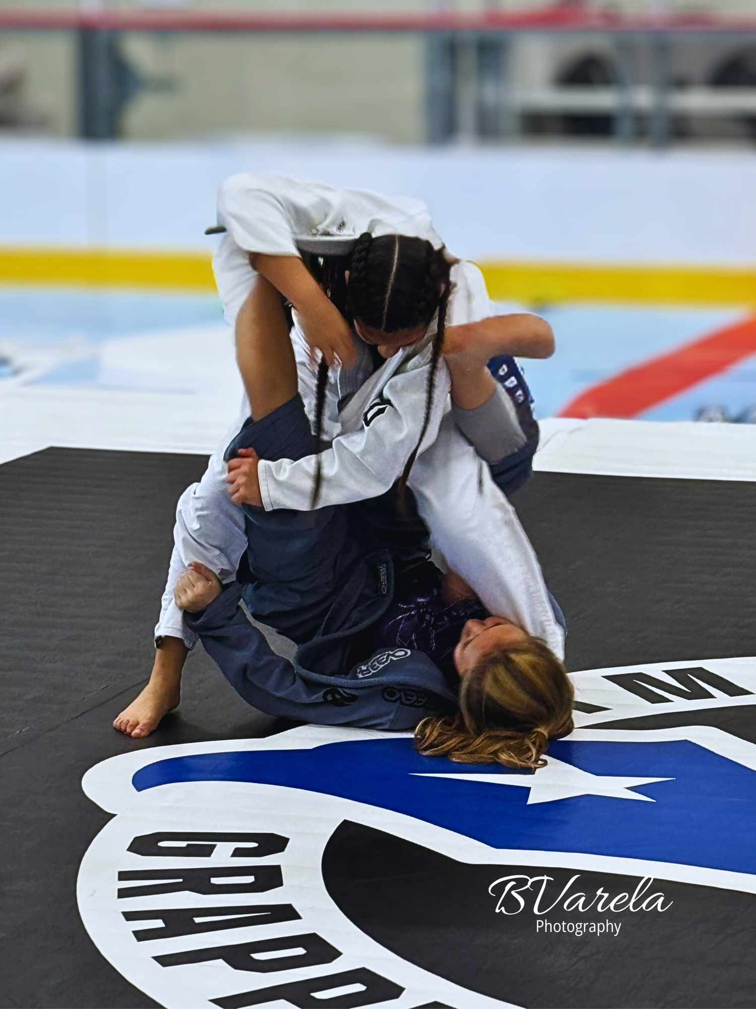 Two female grapplers competing in a jiu-jitsu match on a branded competition mat. sport photographer