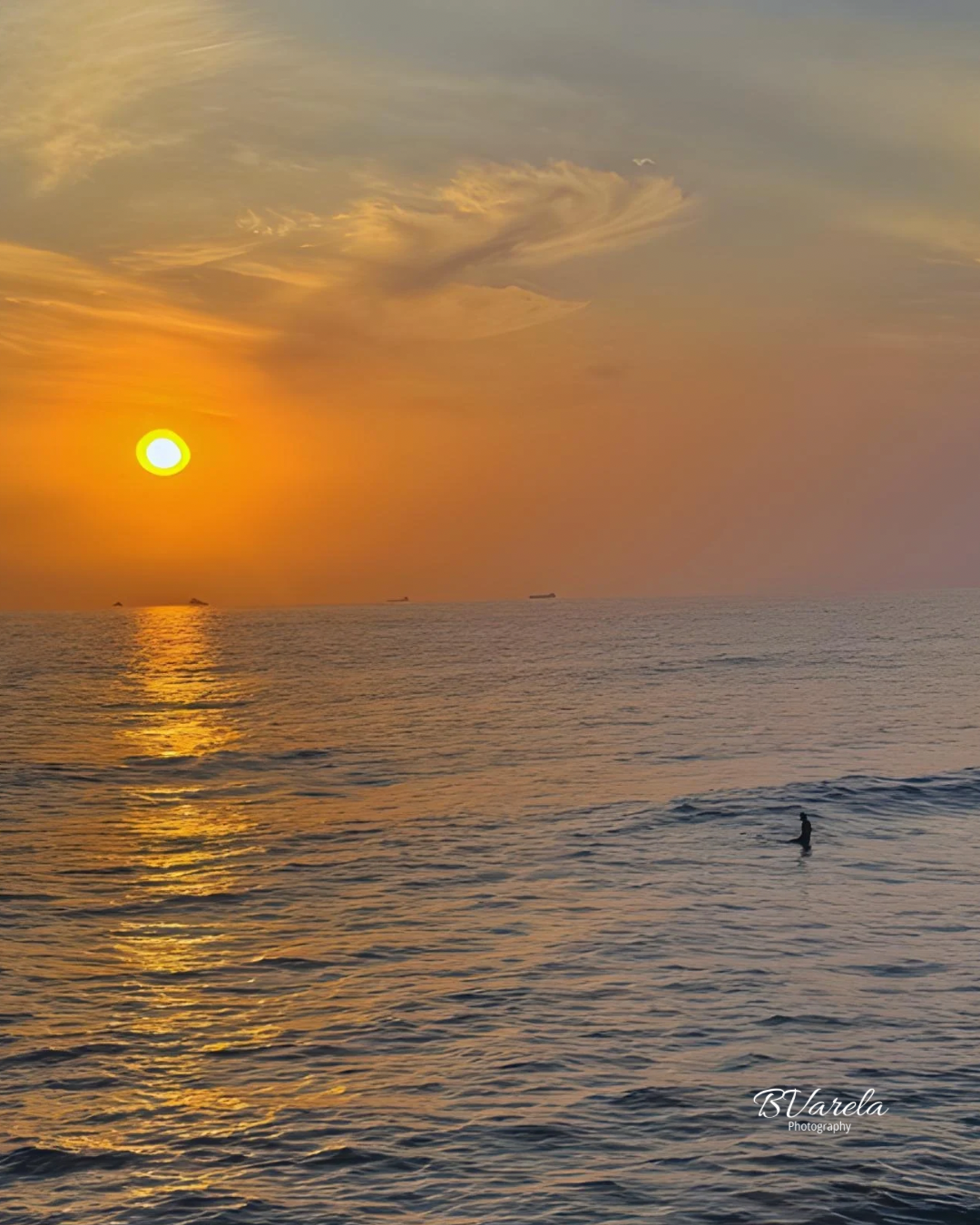 A lone surfer silhouetted against a vibrant orange sunset over calm ocean waters with golden reflections.