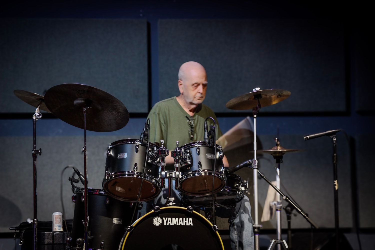 Bald man playing Yamaha drum kit on stage with cymbals in dimly lit performance venue.