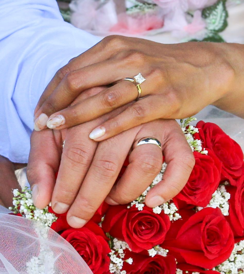 Newlywed couple holding hands over red roses bouquet, wearing wedding rings, symbolizing love and marriage.