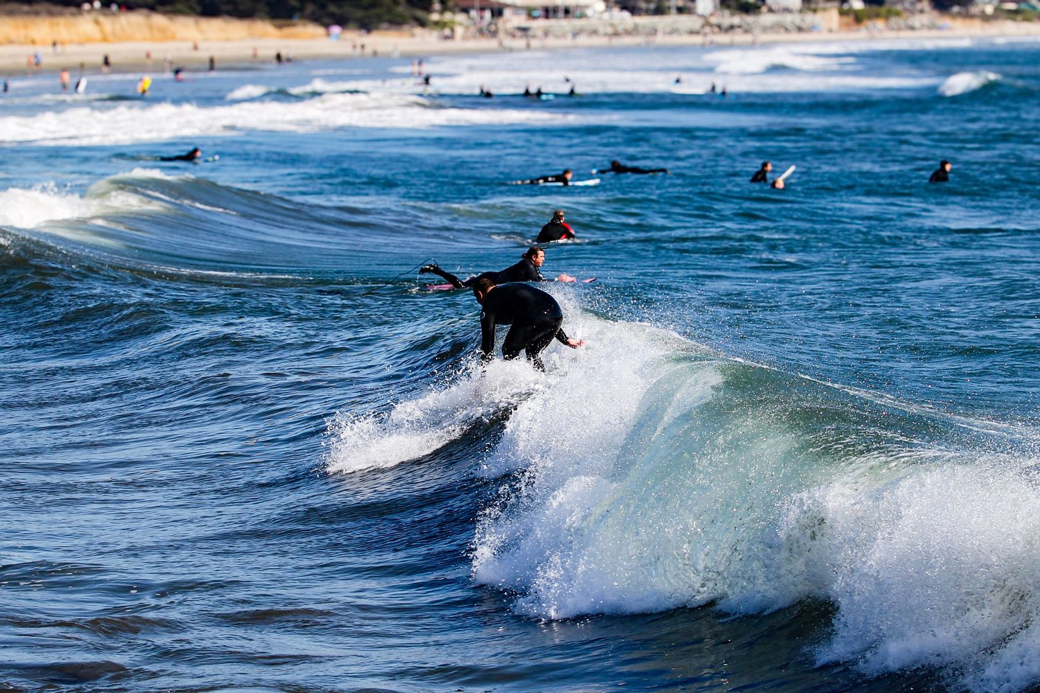 Surfer riding a wave in black wetsuit at a busy beach with other surfers visible in the background.