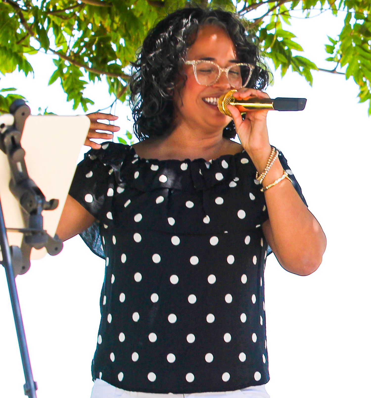 Woman in polka dot blouse singing into gold microphone at outdoor music performance.