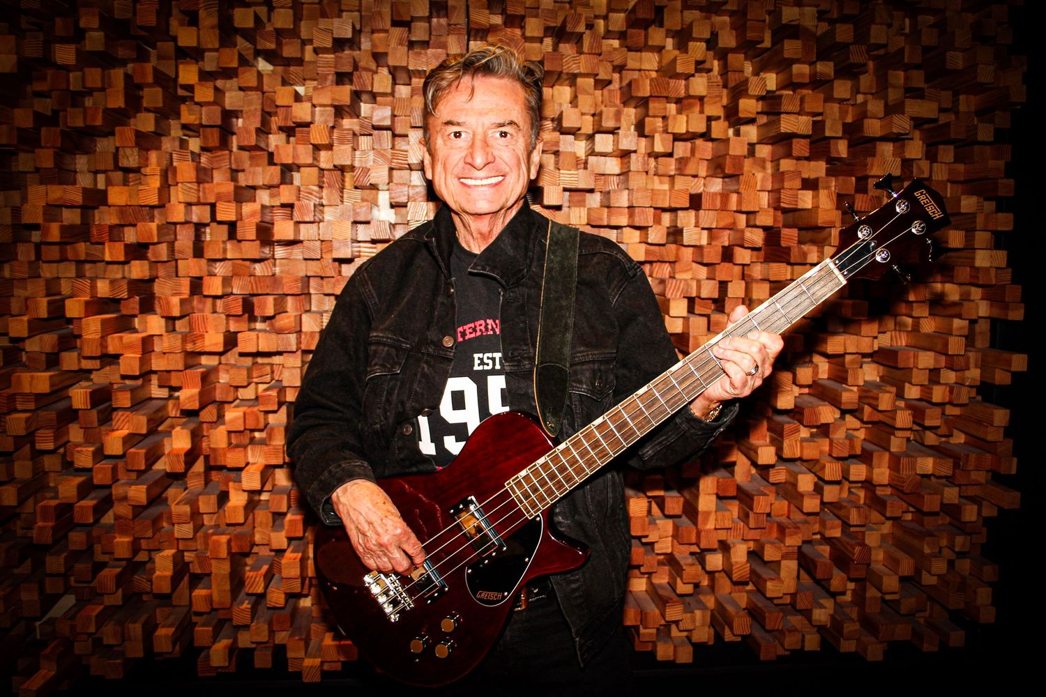 Smiling man holding a dark electric guitar against a wooden block wall backdrop.