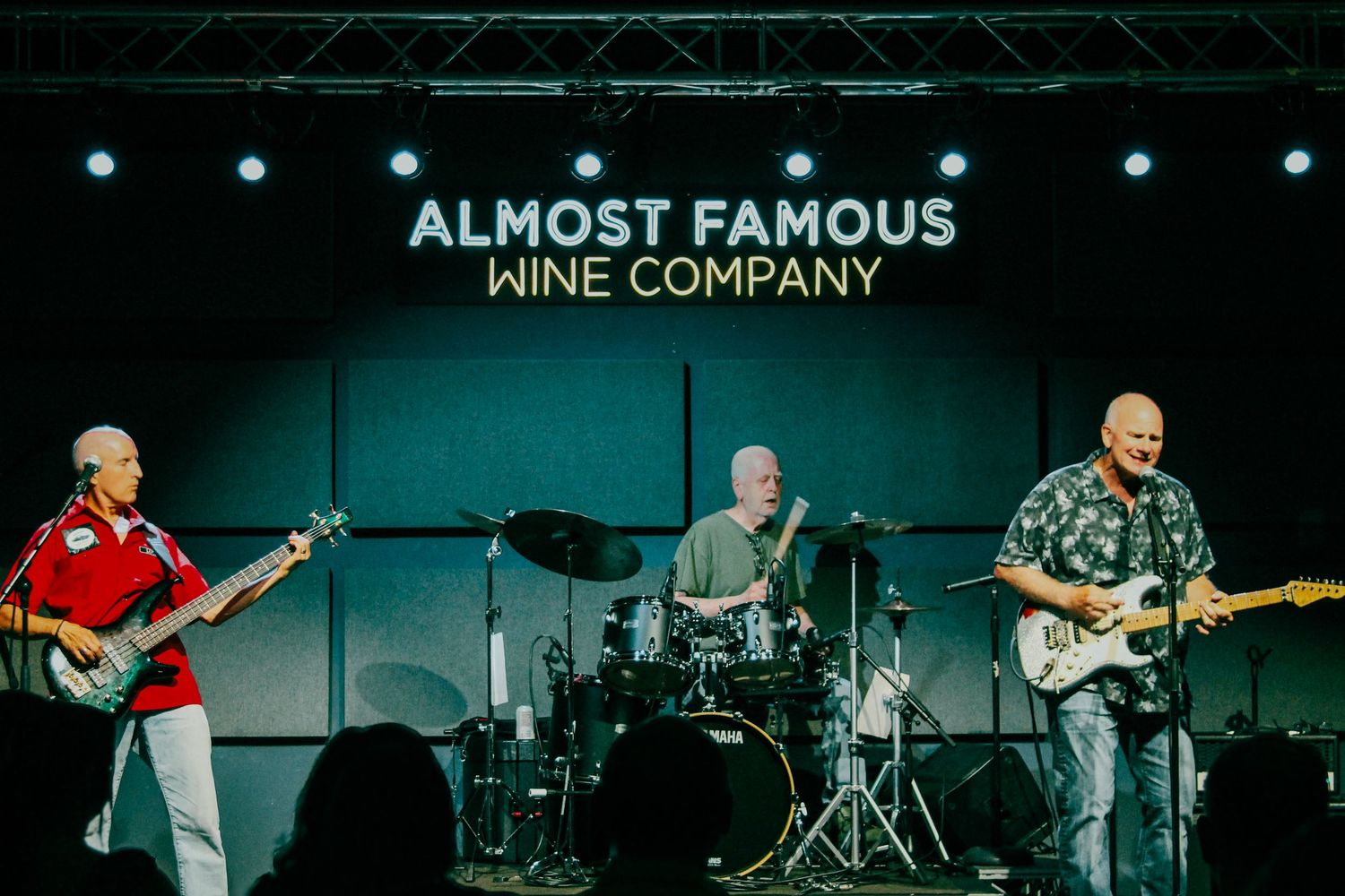 Three musicians perform live on stage at Almost Famous Wine Company, featuring guitar and drums.