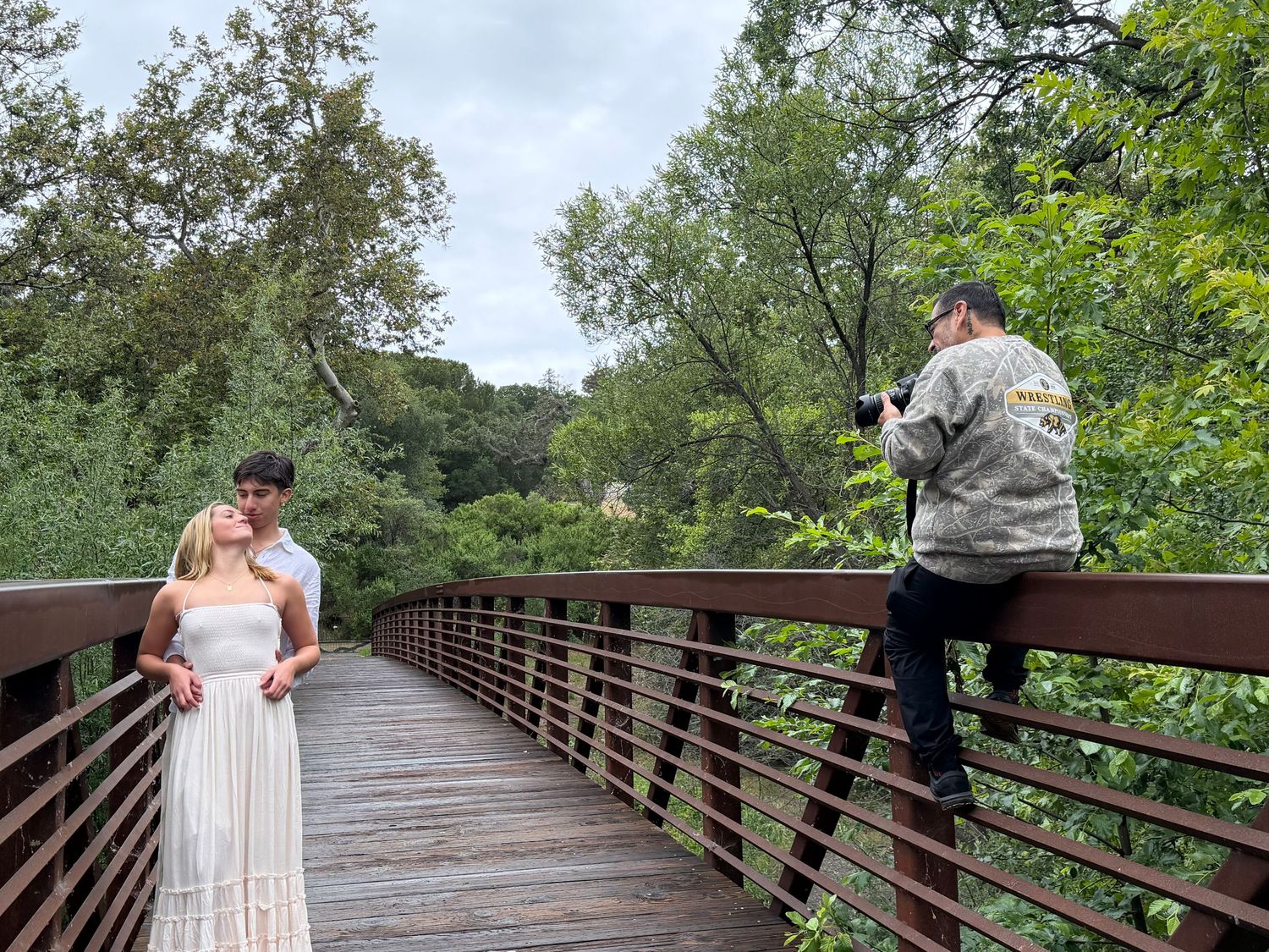 Photographer balancing on bridge railing captures couple posing on wooden footbridge surrounded by lush green trees.
