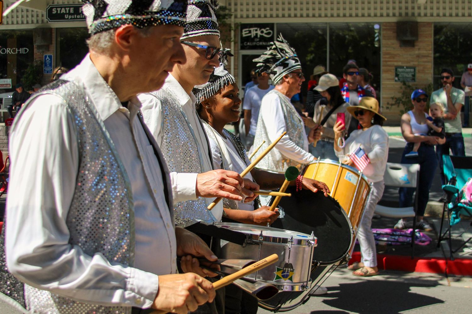 Musicians in silver sequined costumes play drums during a festive outdoor parade on a sunny day.