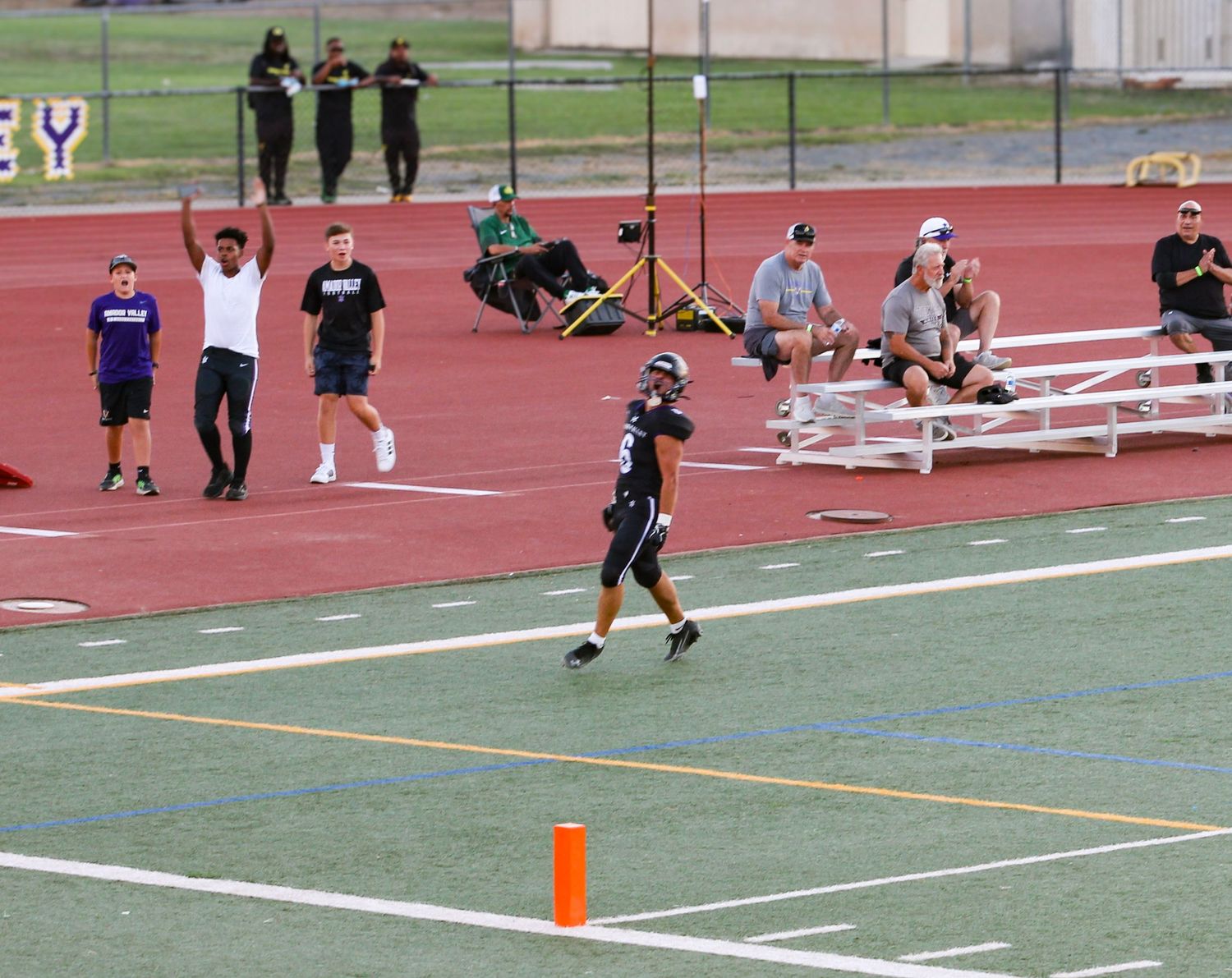 Football player scores a touchdown in end zone during a game, with coaches and spectators watching from the sidelines.