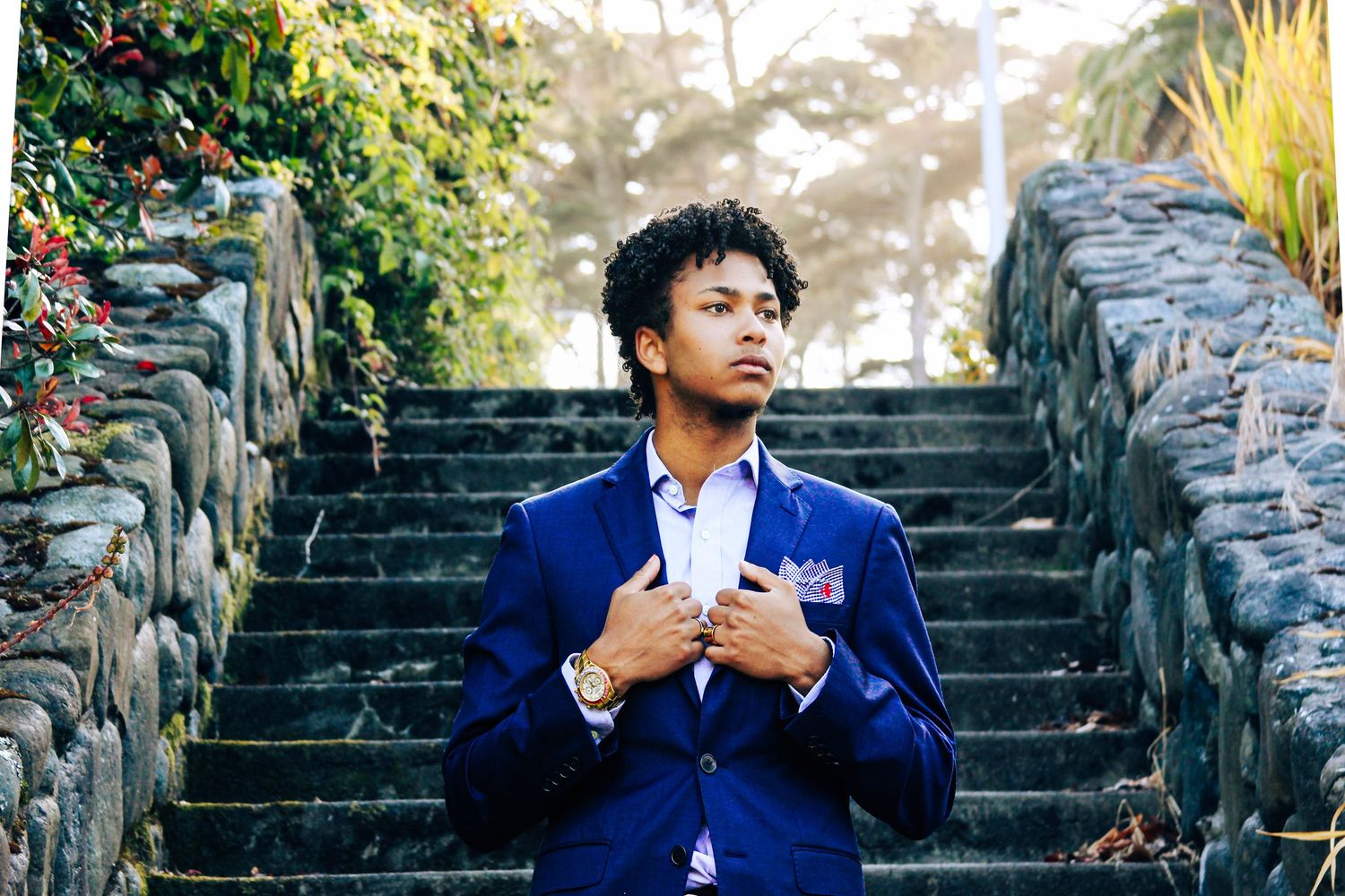 Young man in blue blazer with pocket square adjusts jacket on stone stairway outdoors.