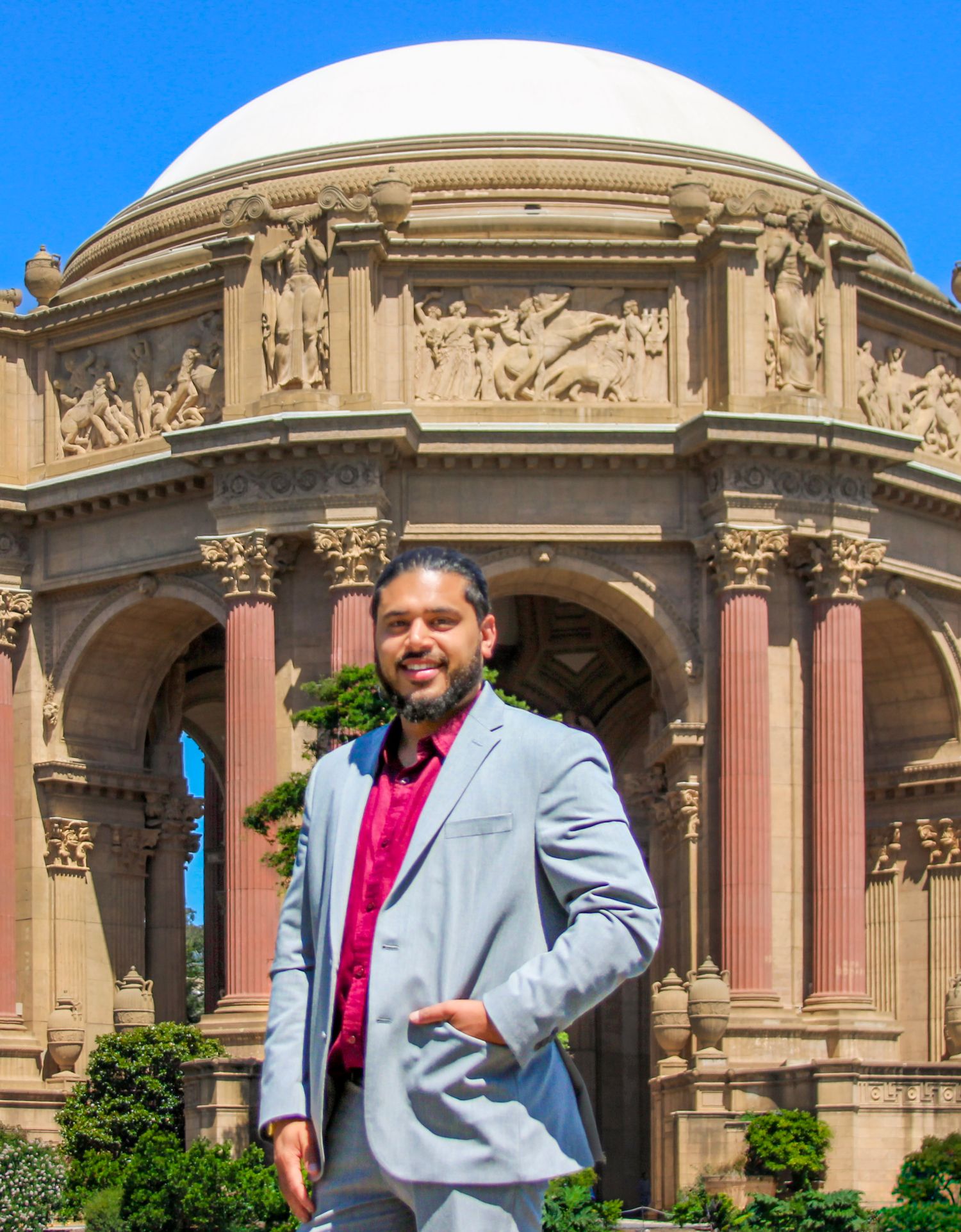 Man in gray suit poses before the Palace of Fine Arts rotunda in San Francisco on a sunny day.