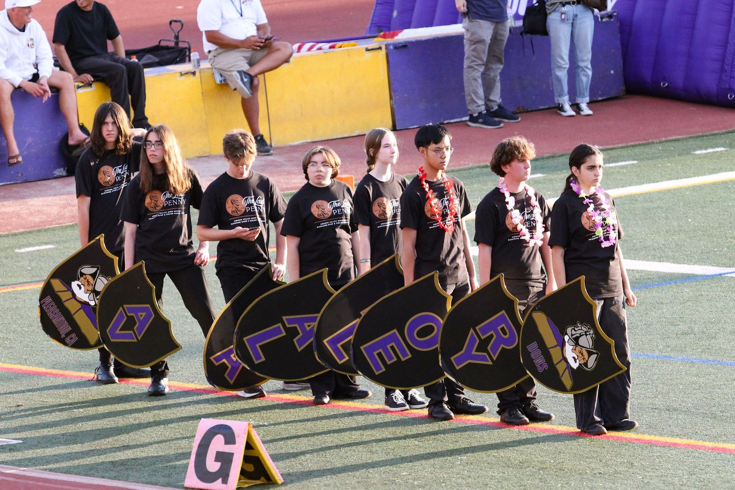 Marching band members in black uniforms carry decorative shields on a football field during a performance.