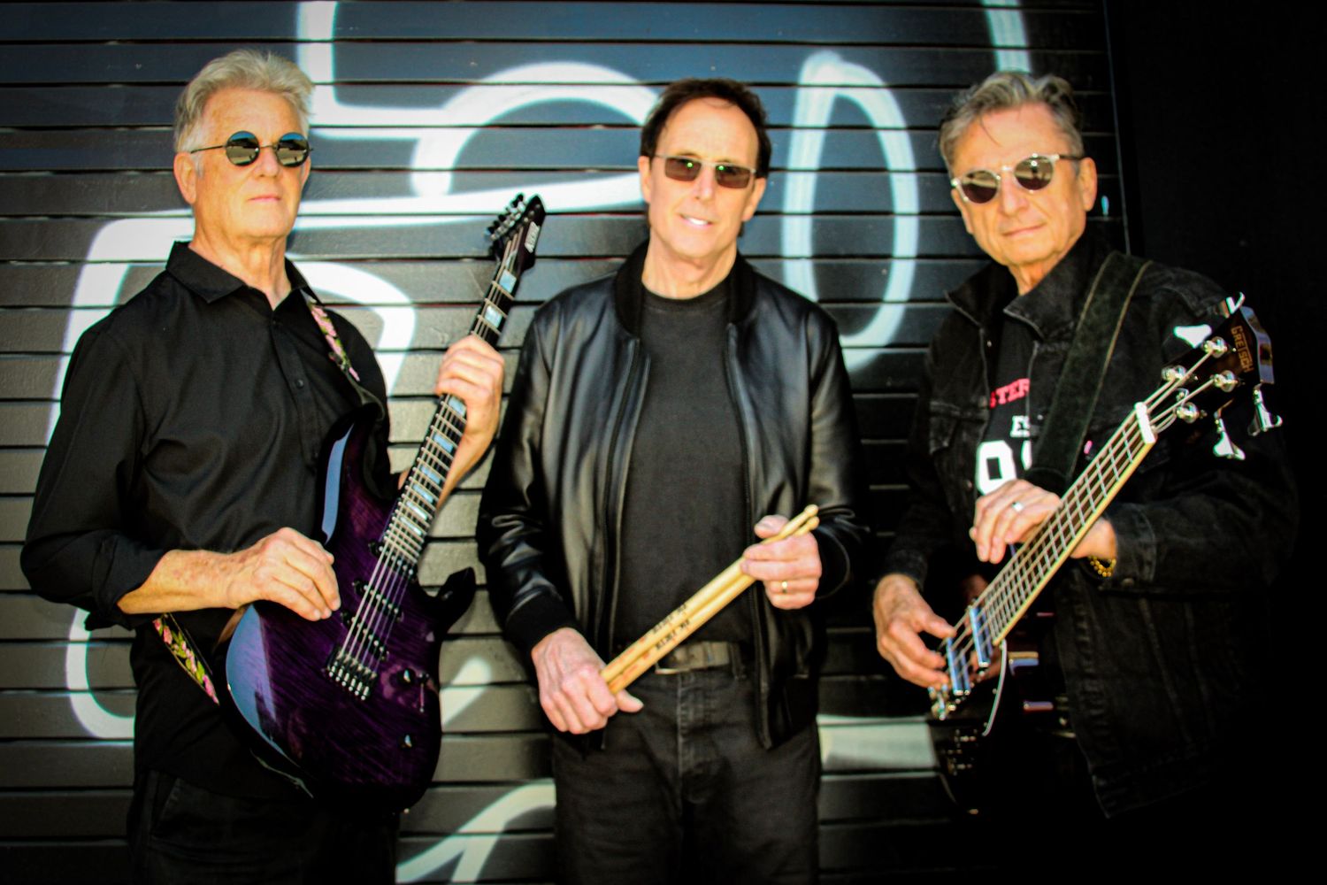 Three male musicians posing with electric guitars against a dark graffiti wall in matching black outfits.