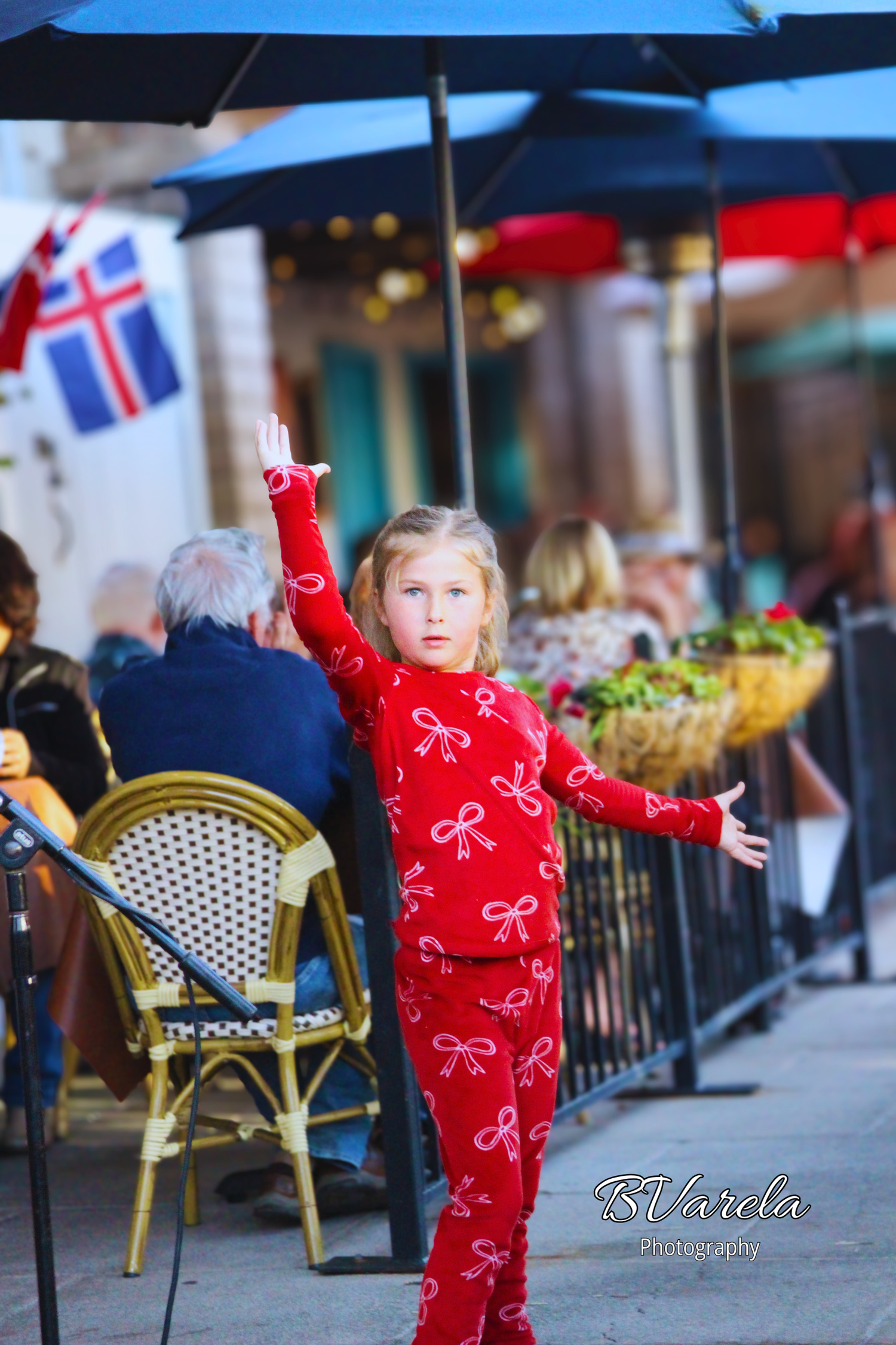 Young girl in red bow pajamas poses with arms outstretched at outdoor cafe patio with blue umbrellas.
