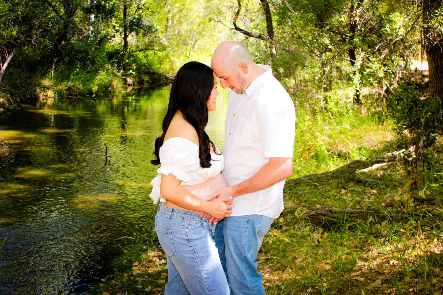 Expecting couple sharing an intimate moment by a serene green creek, dressed in white tops and jeans.