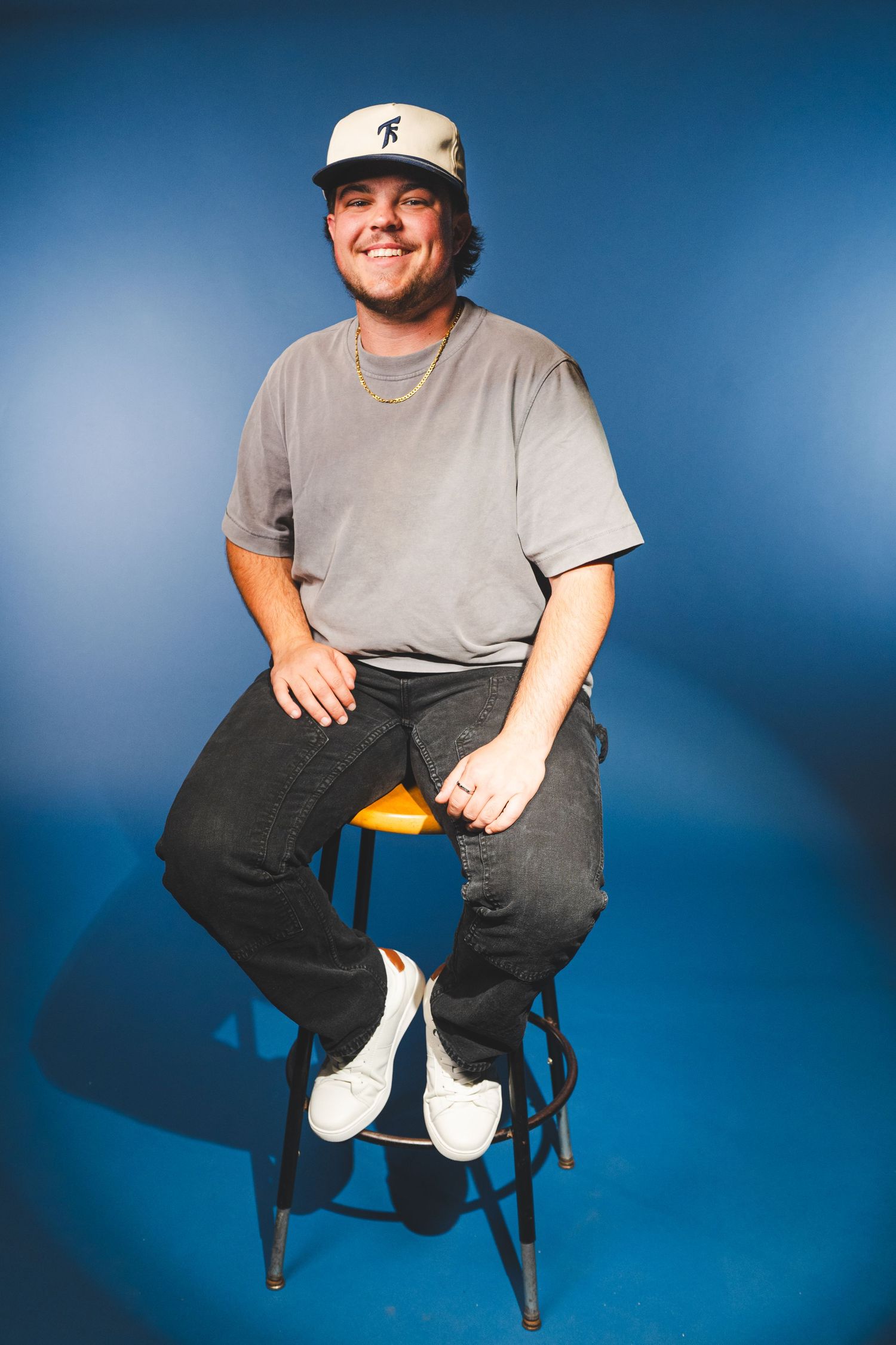 Young man in gray tee and cap sits smiling on a stool against a blue studio background.