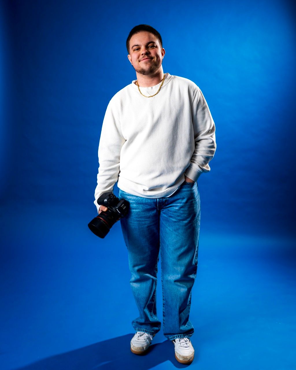Young man in white sweatshirt and jeans holds a camera, posing confidently against a vibrant blue studio backdrop.