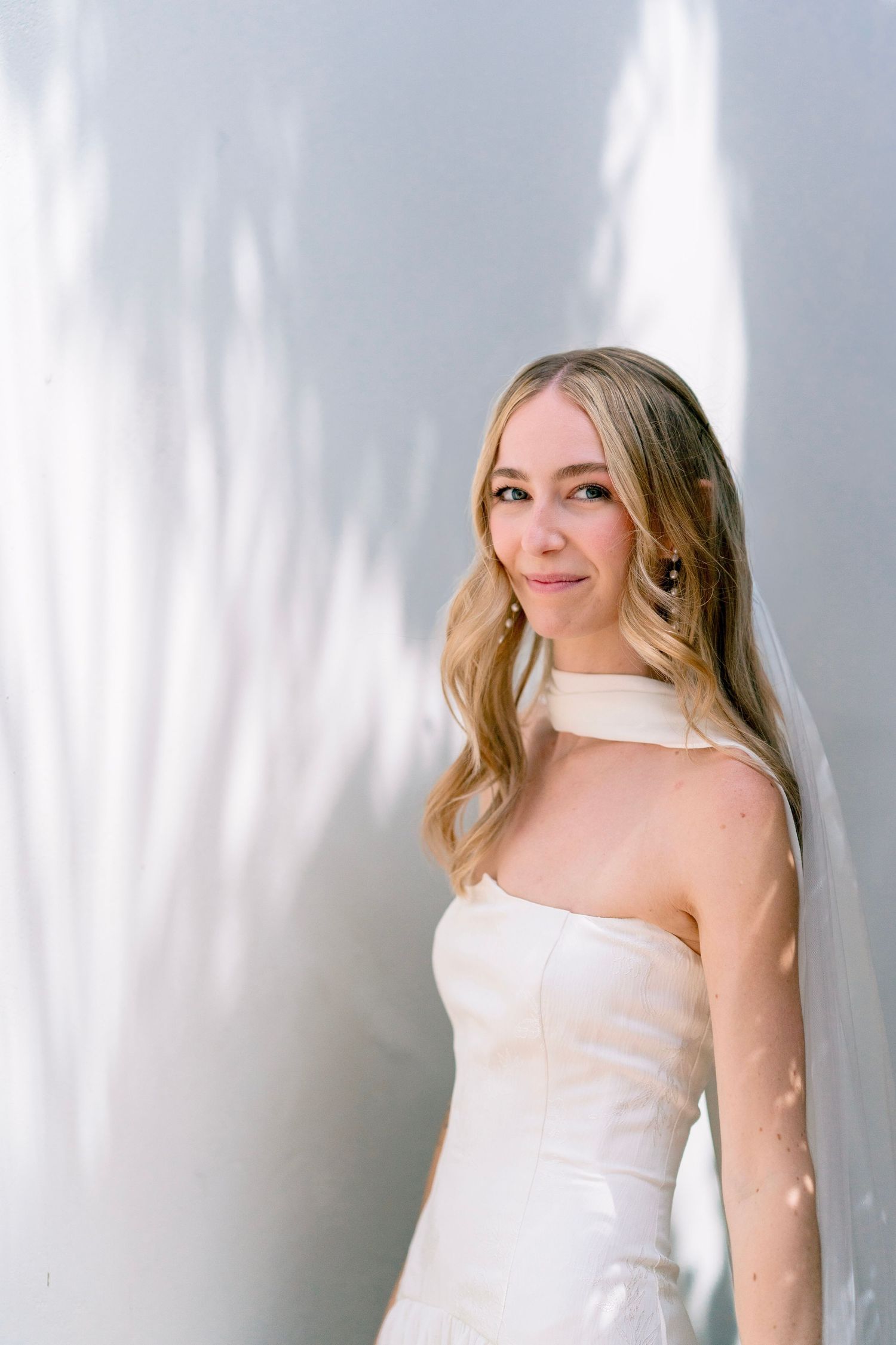 Elegant bride in strapless white gown and veil poses against a light gray wall with soft leaf shadows.