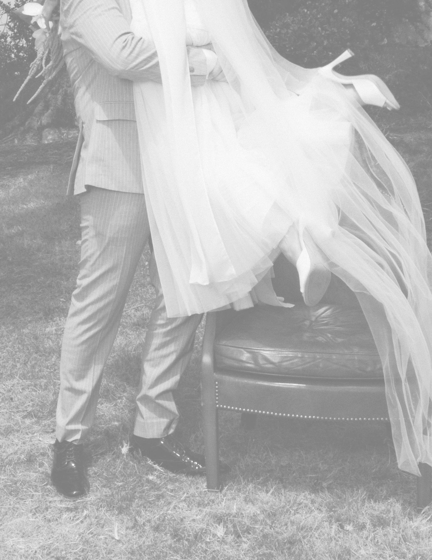 Black and white photo of a bride and groom standing outdoors, her flowing veil billowing beside a leather chair.