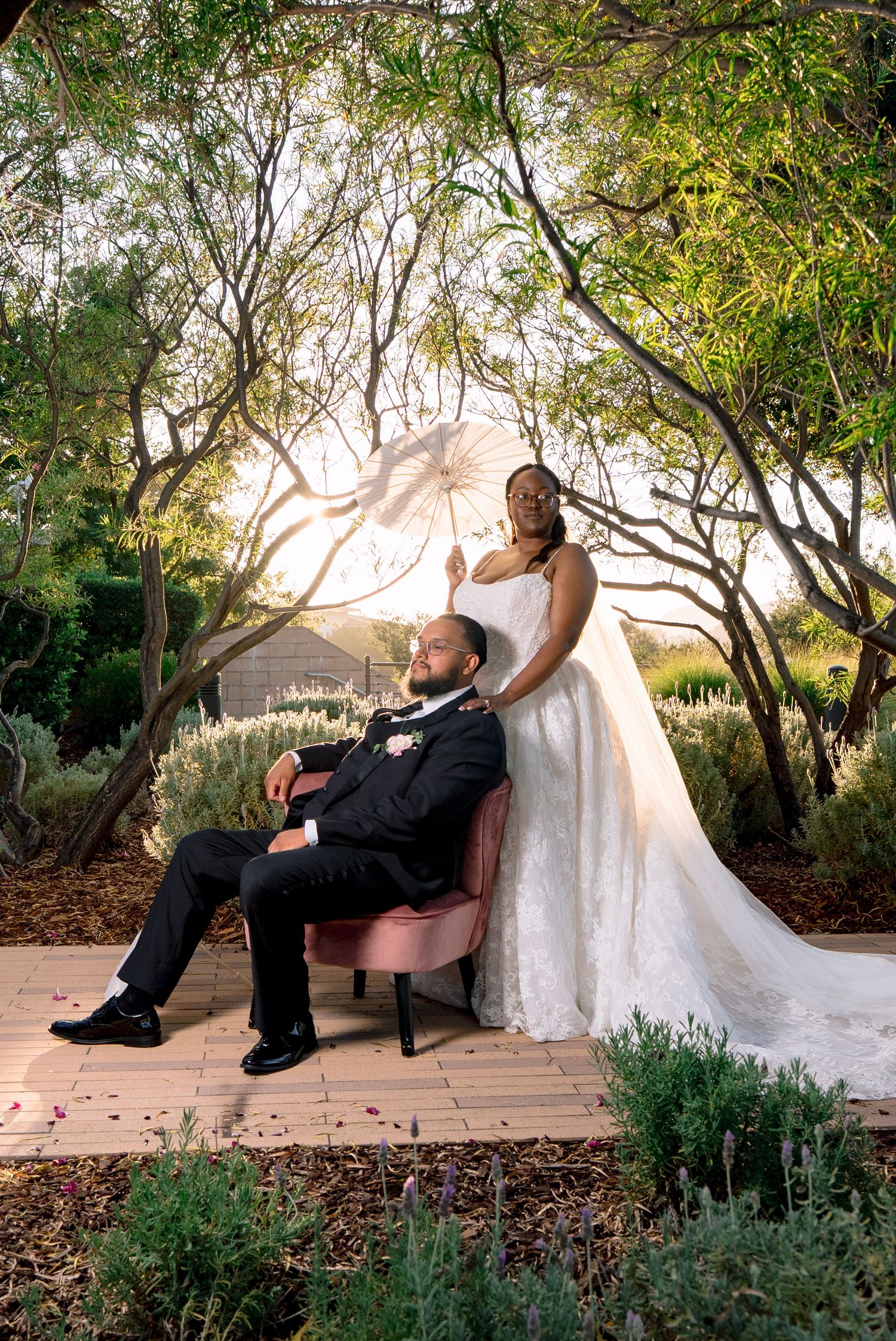 Bride holding white parasol stands behind groom seated in pink chair, surrounded by lush garden trees at golden hour.