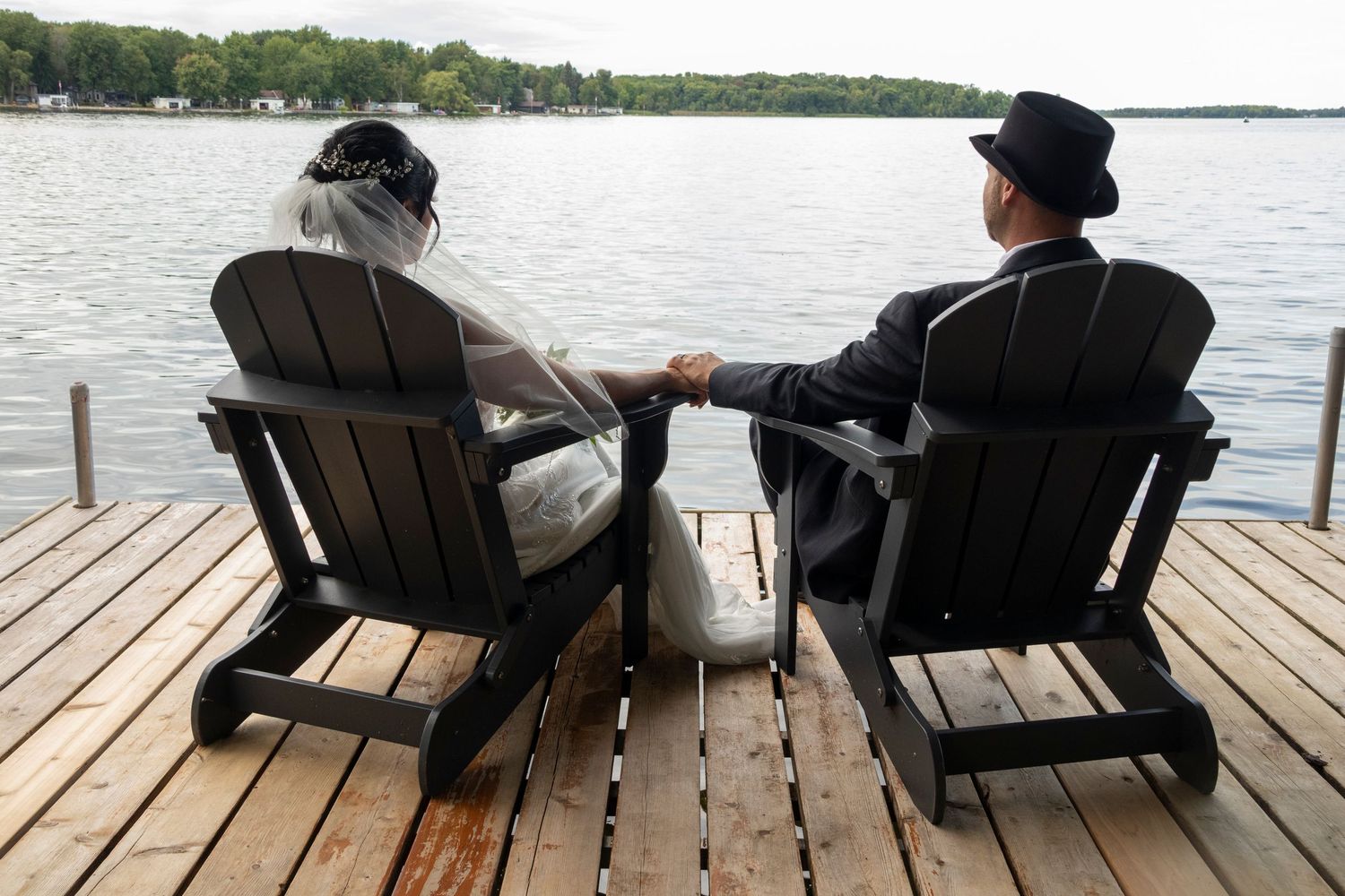 Bride and groom sitting in Adirondack chairs looking out at the lake holding hands
