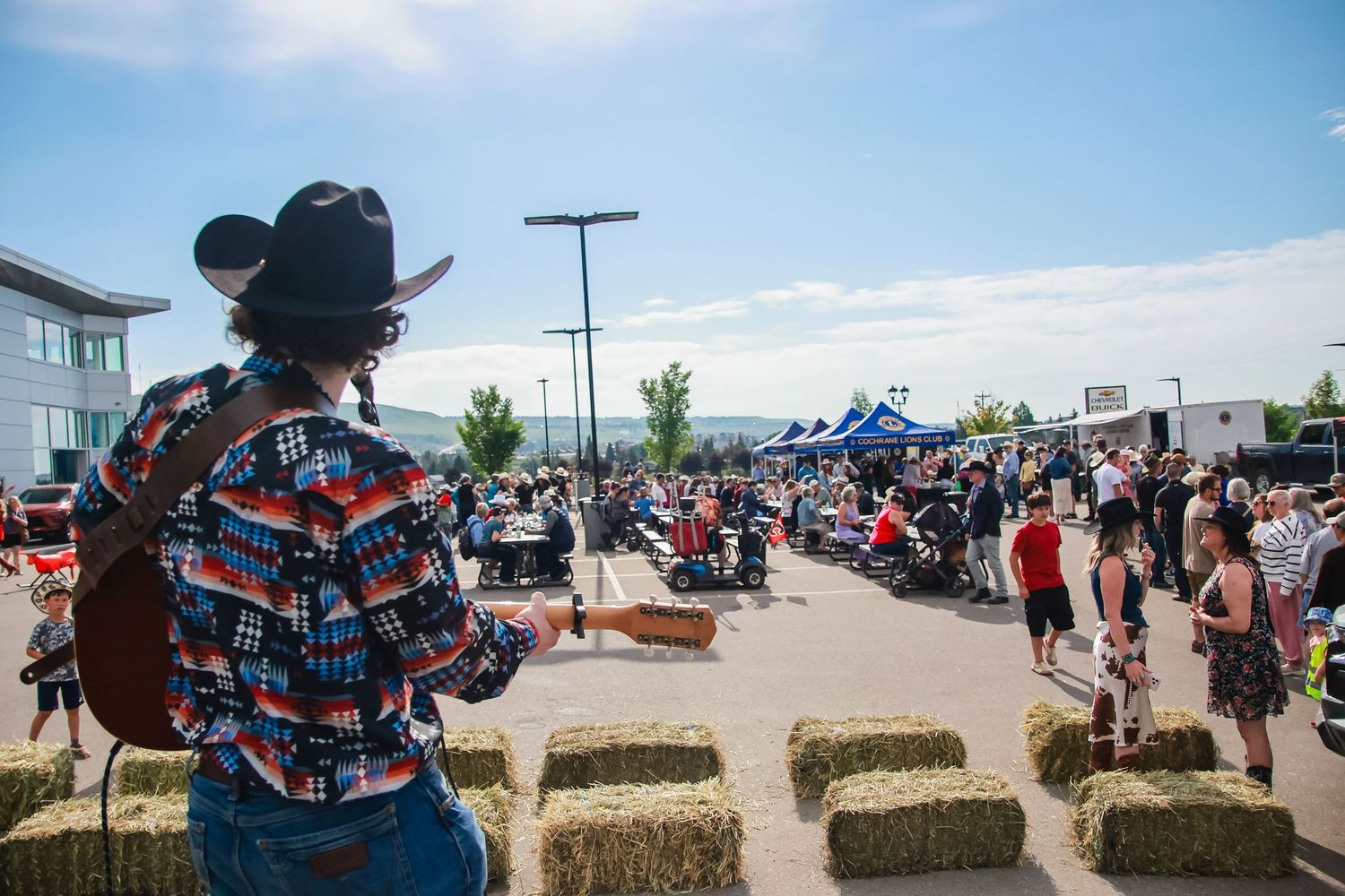 A guitarist in a cowboy hat overlooks a lively outdoor country festival with hay bales and blue tents.